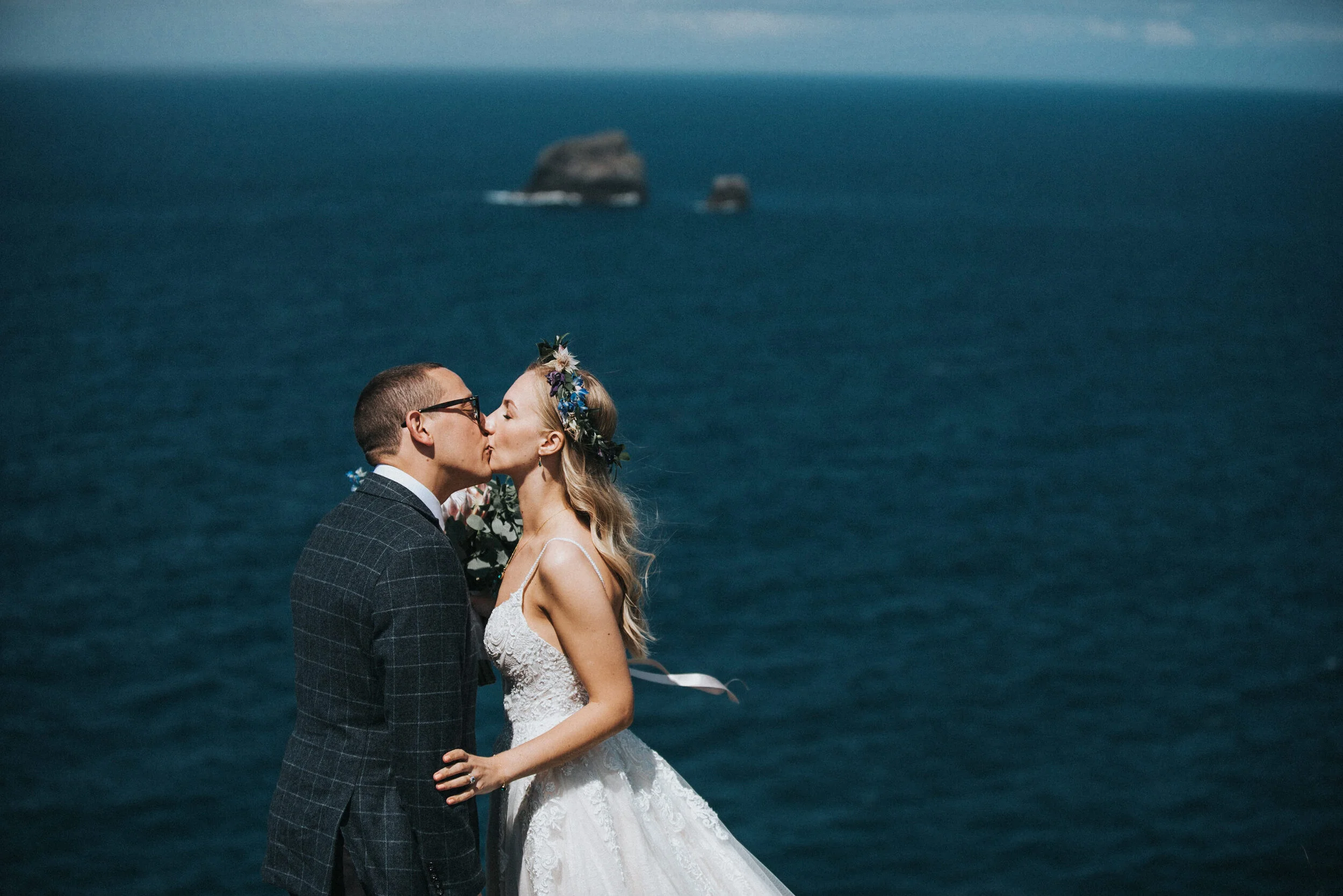 A bride and groom kiss by the ocean during their wedding in Cornwall, with rocky islands in the background.