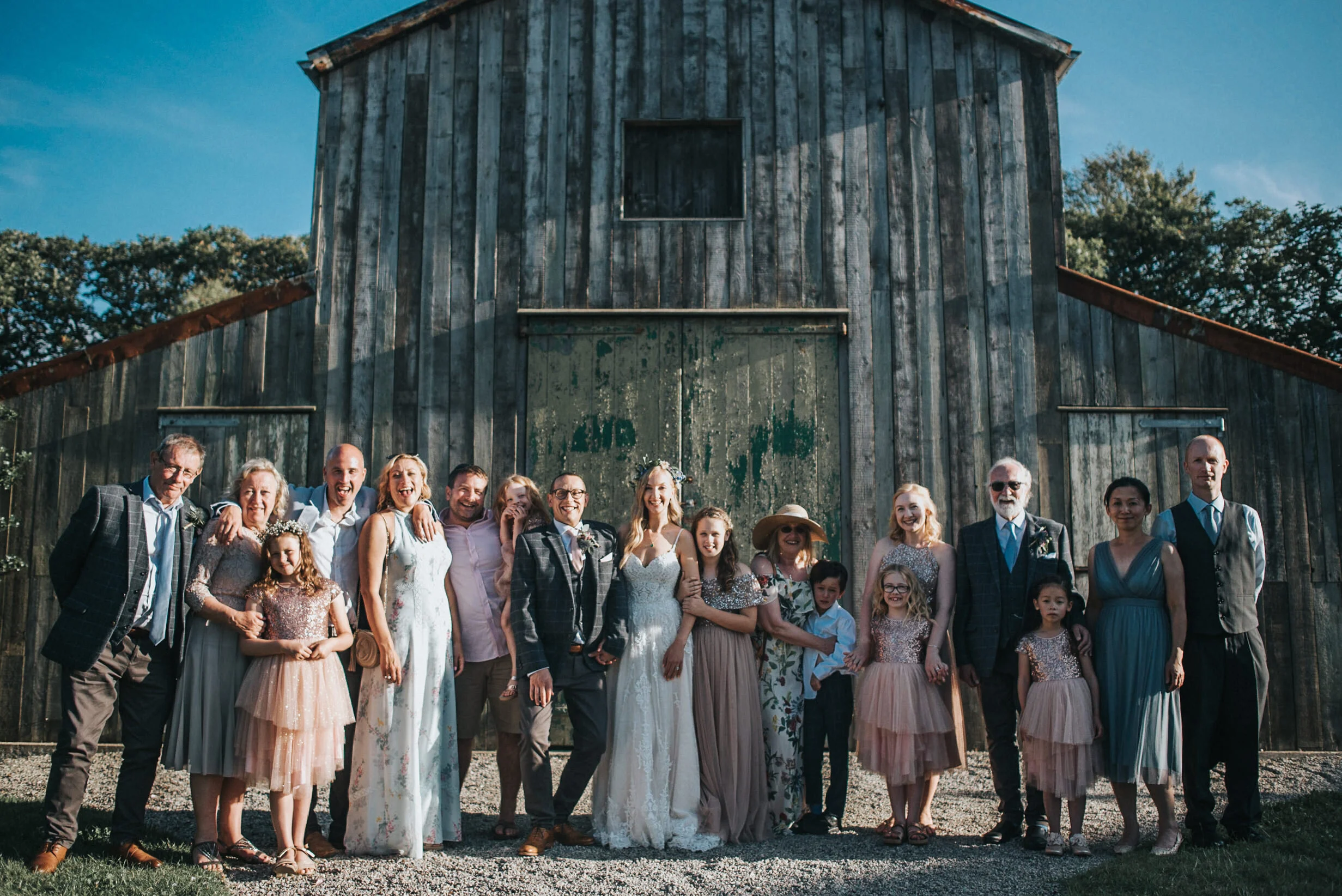 A large group of people, including adults and children, gathered outdoors for a wedding photo in front of a rustic wooden barn on a sunny day.