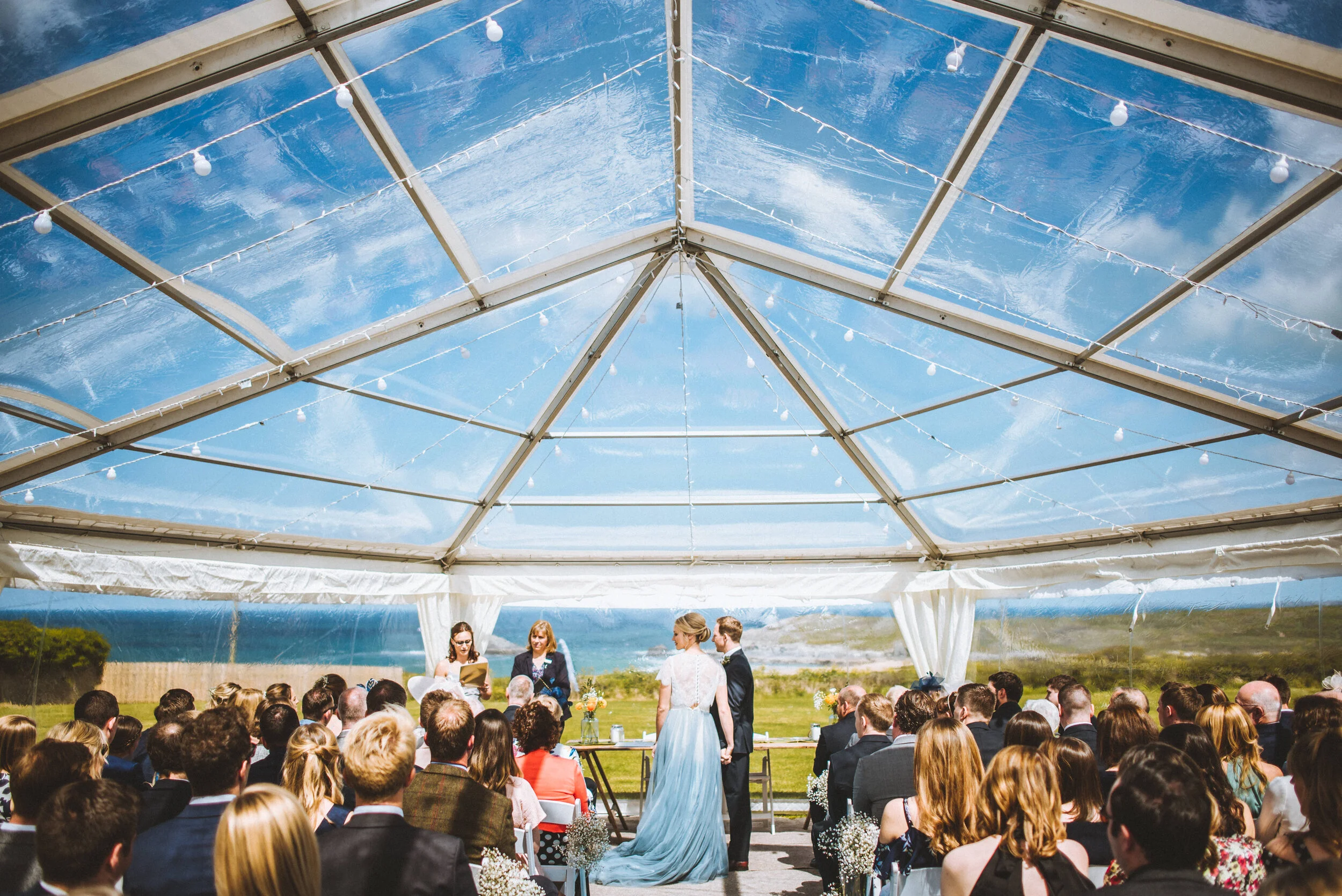 Wedding ceremony taking place under a glass tent with string lights, overlooking a scenic ocean view, with bride and groom facing officiant, surrounded by seated guests.