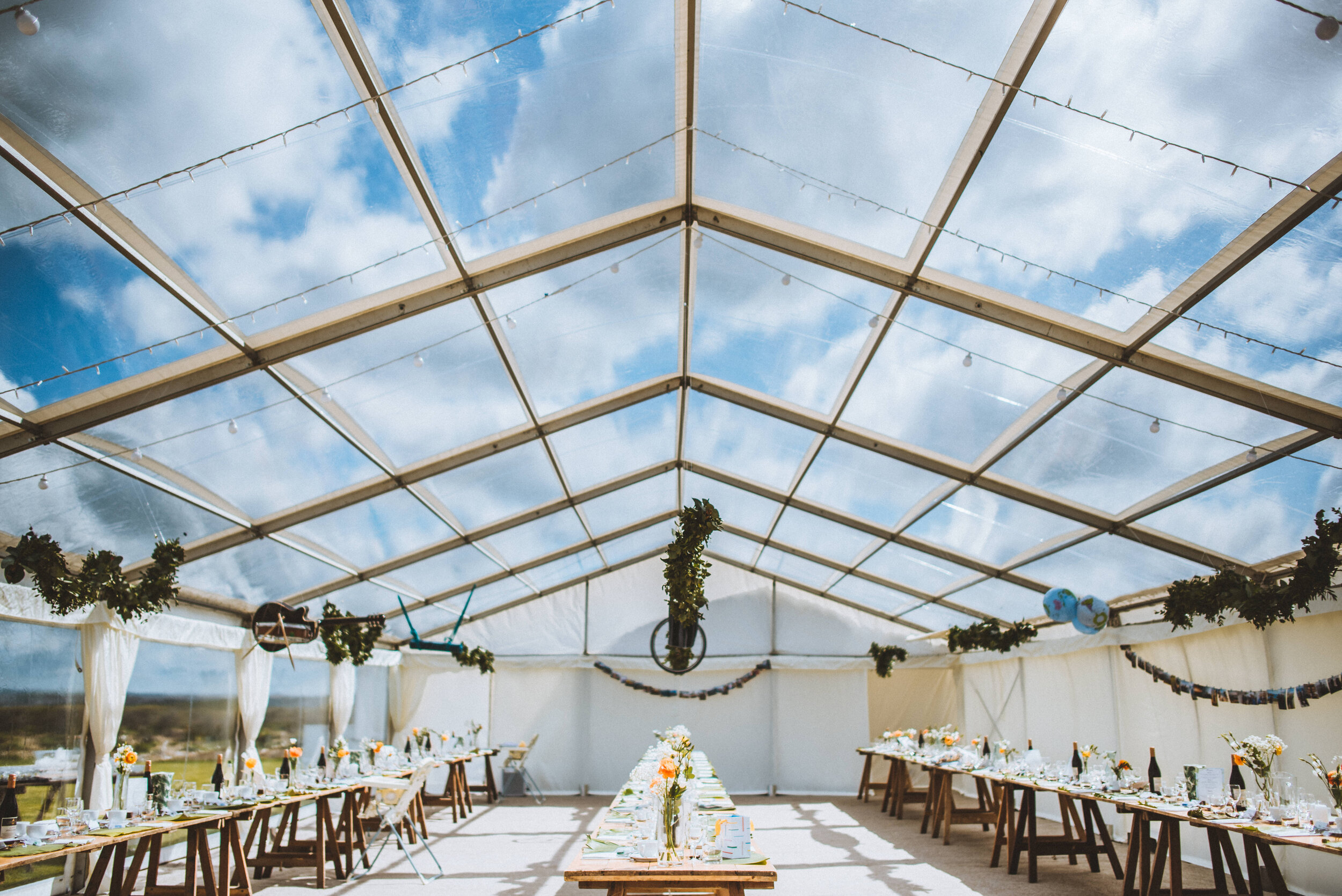 Indoor event space with long dining tables, decorated with flowers and tableware, under a transparent roof showing a blue sky with clouds, illuminated with string lights.