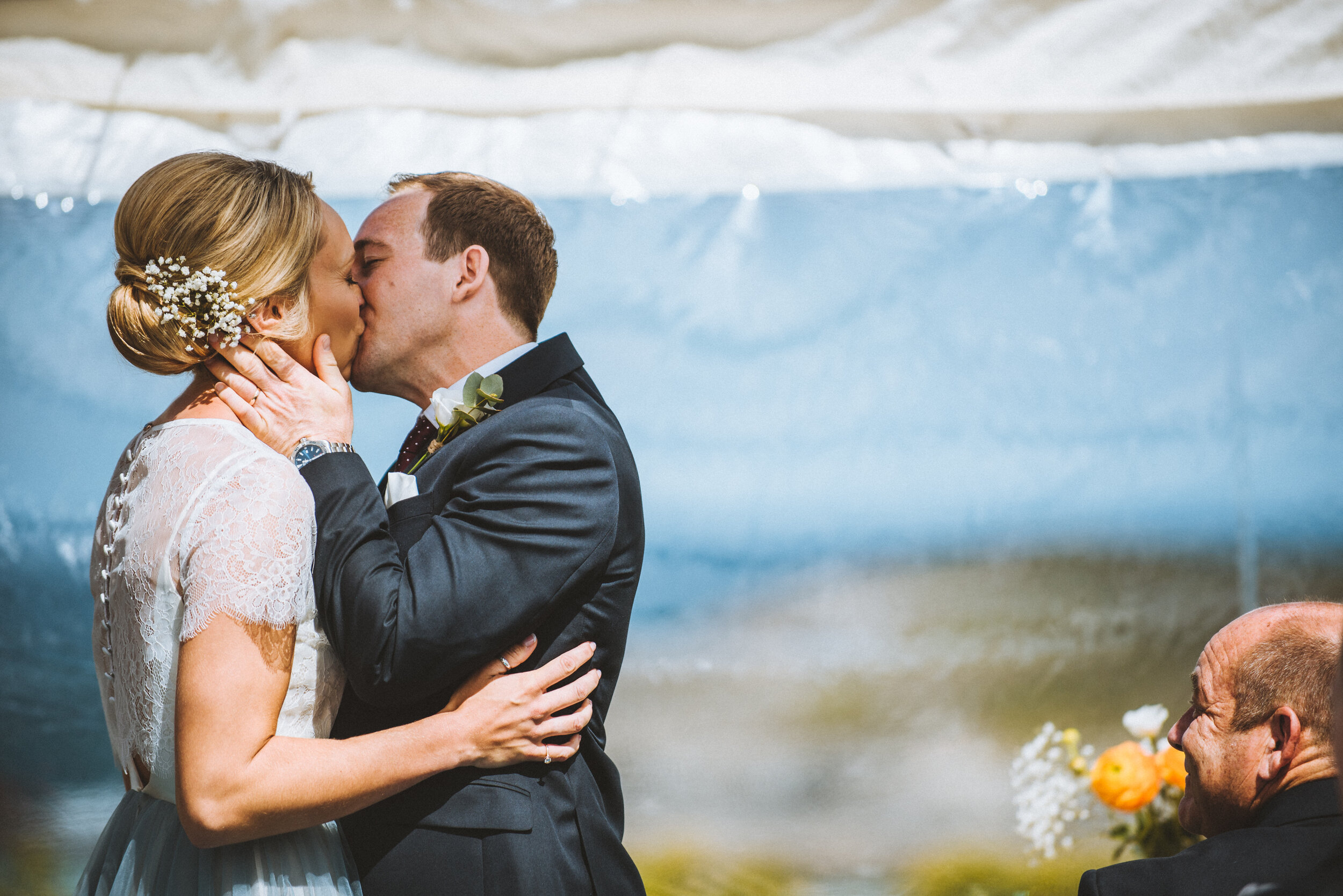A bride and groom kissing during their wedding ceremony, with a man in black formal attire smiling and looking on in the background, and a bouquet of flowers on a table nearby.