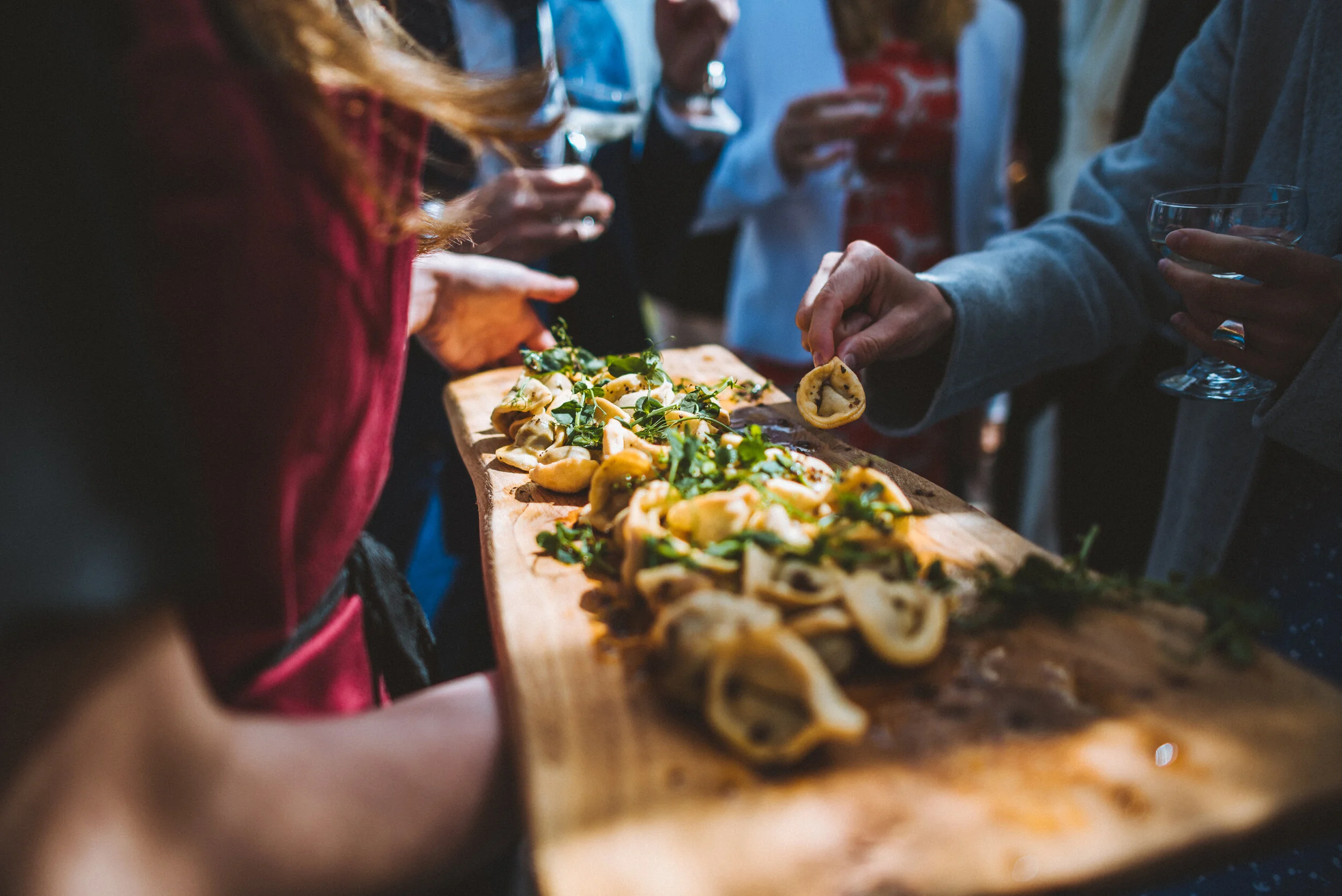 People gathered around a wooden serving board with pasta and greens, serving themselves at a social event.