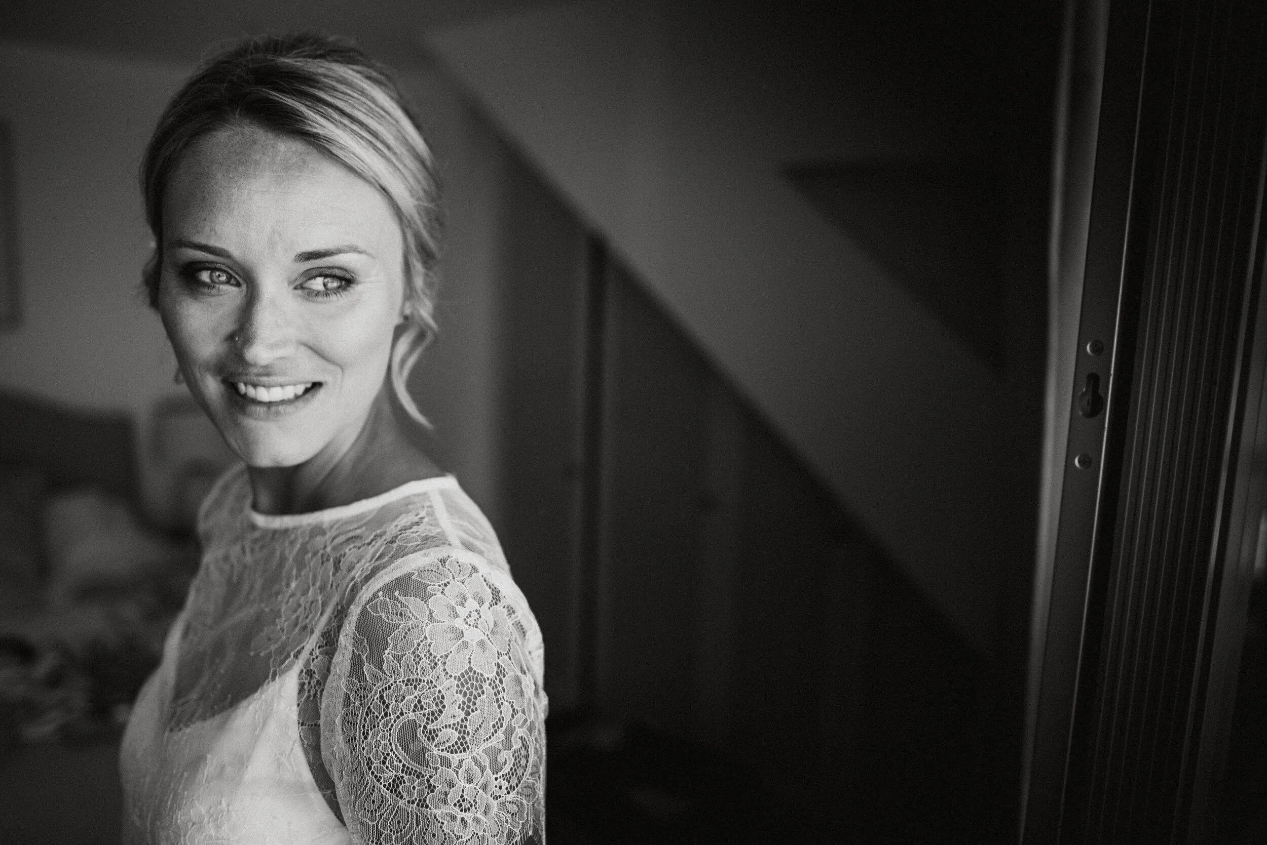 A black and white photo of a smiling woman with short blonde hair, wearing a lace top, looking over her shoulder.
