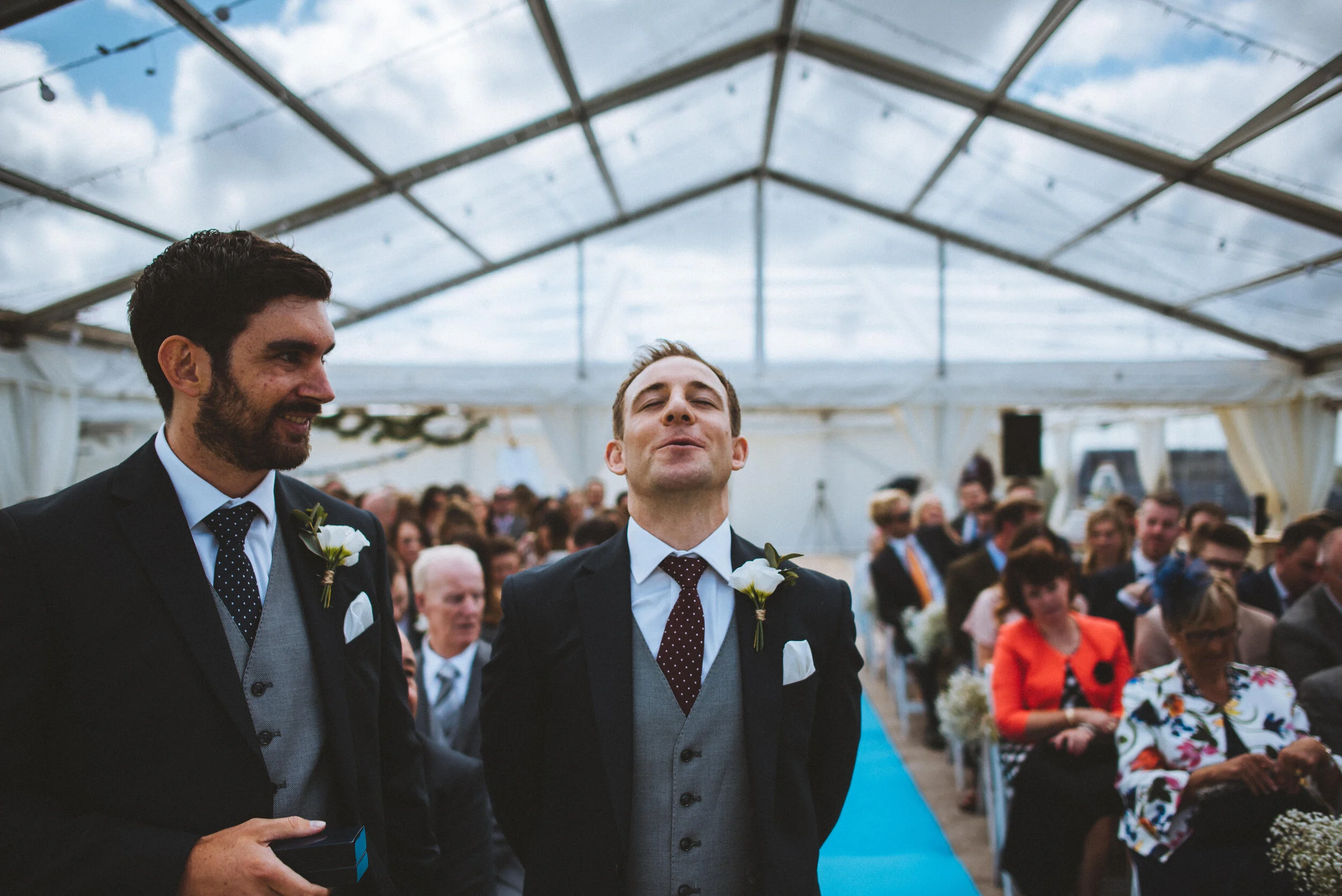 Two men in suits with boutonnieres, one smiling and the other making a face, stand in front of seated wedding guests in a canopy tent with string lights.
