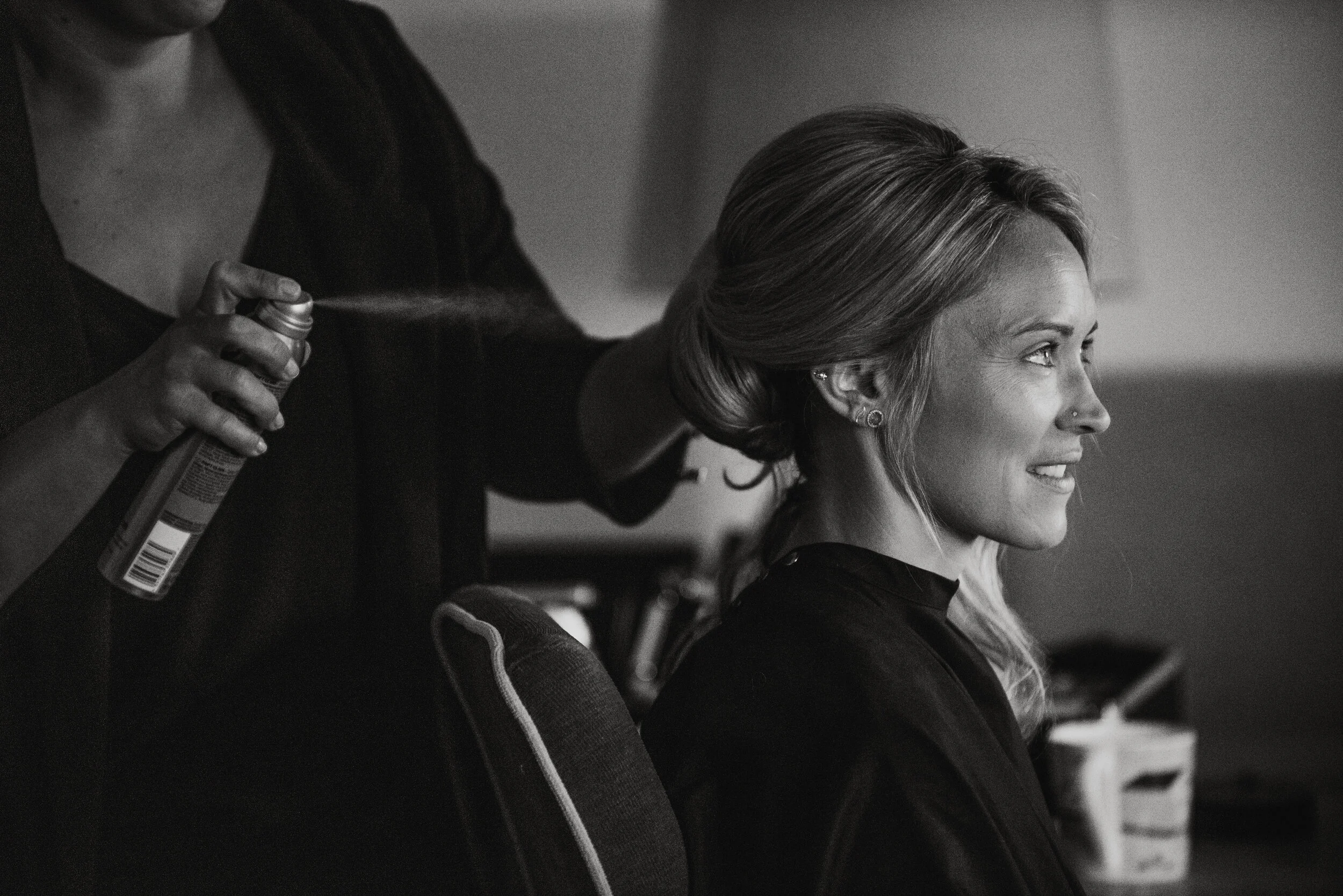 A woman sitting and smiling as her hair is being styled by a hairstylist.