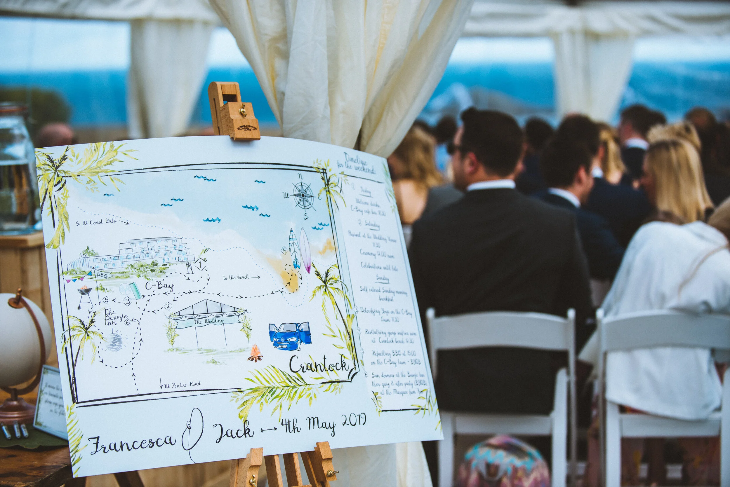 A large wedding map sign displayed on an easel in a ceremony tent, with guests seated in the background.