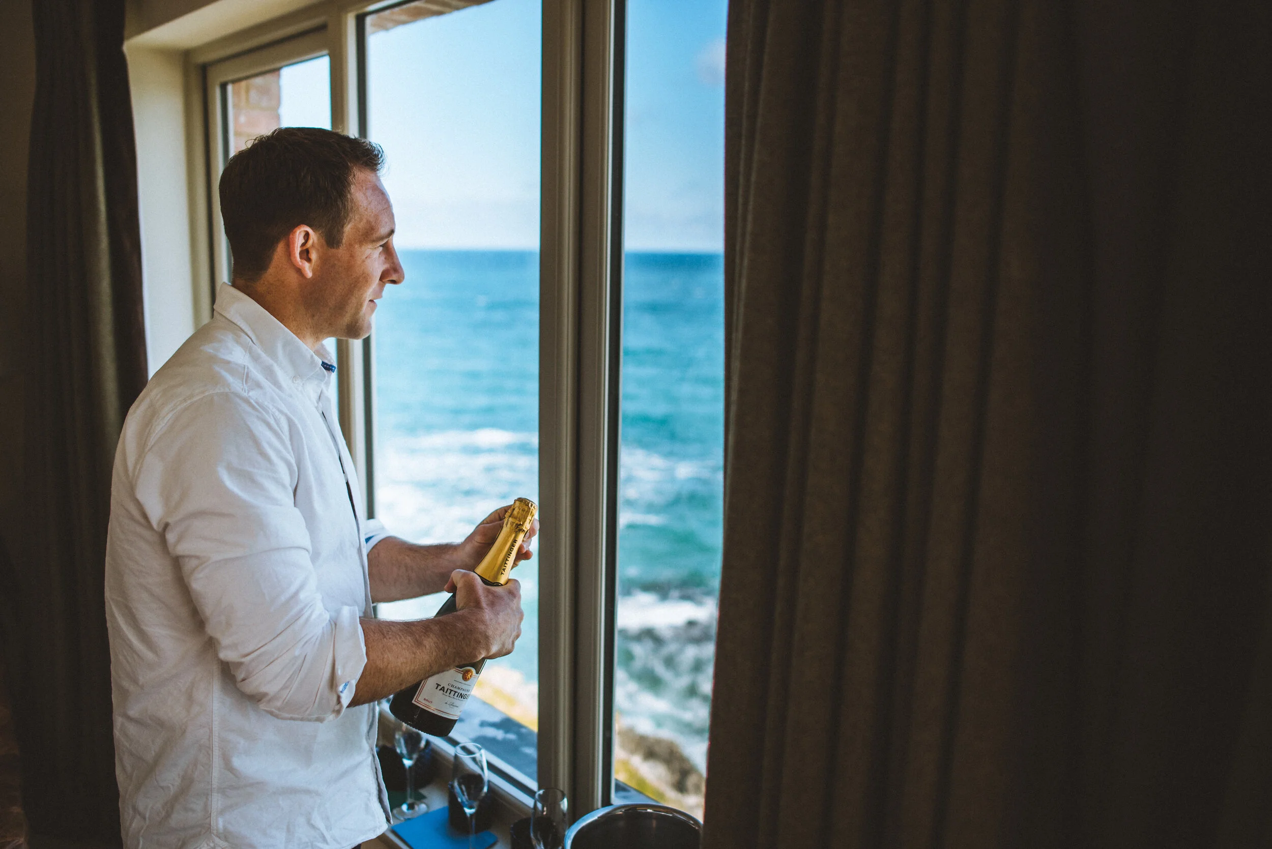 Man in white shirt holding a bottle of champagne near a large window overlooking the ocean.