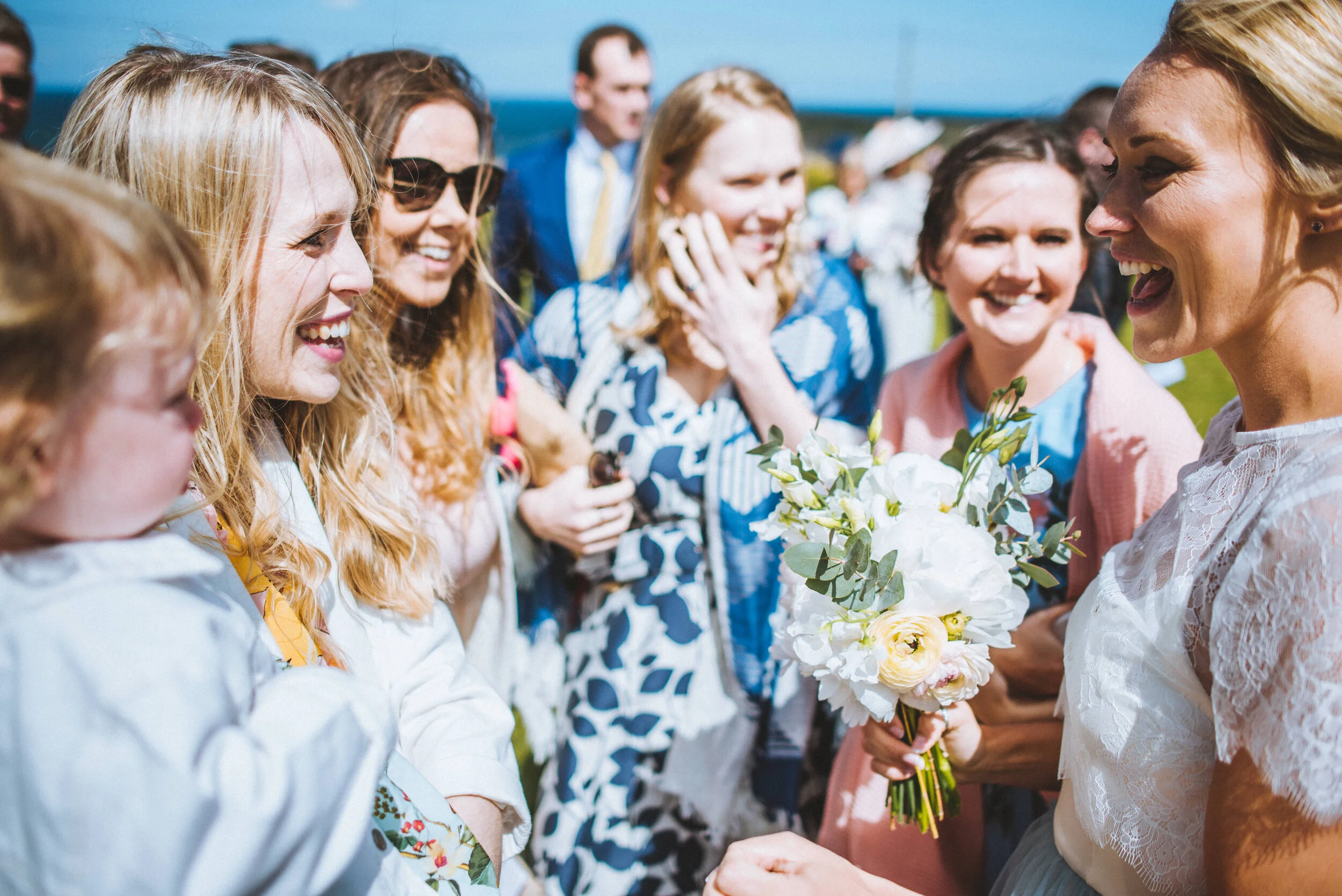 A woman in a white lace dress holding a bouquet of white and yellow flowers is smiling and talking to a group of women and men outdoors on a sunny day, with the ocean or lake in the background.