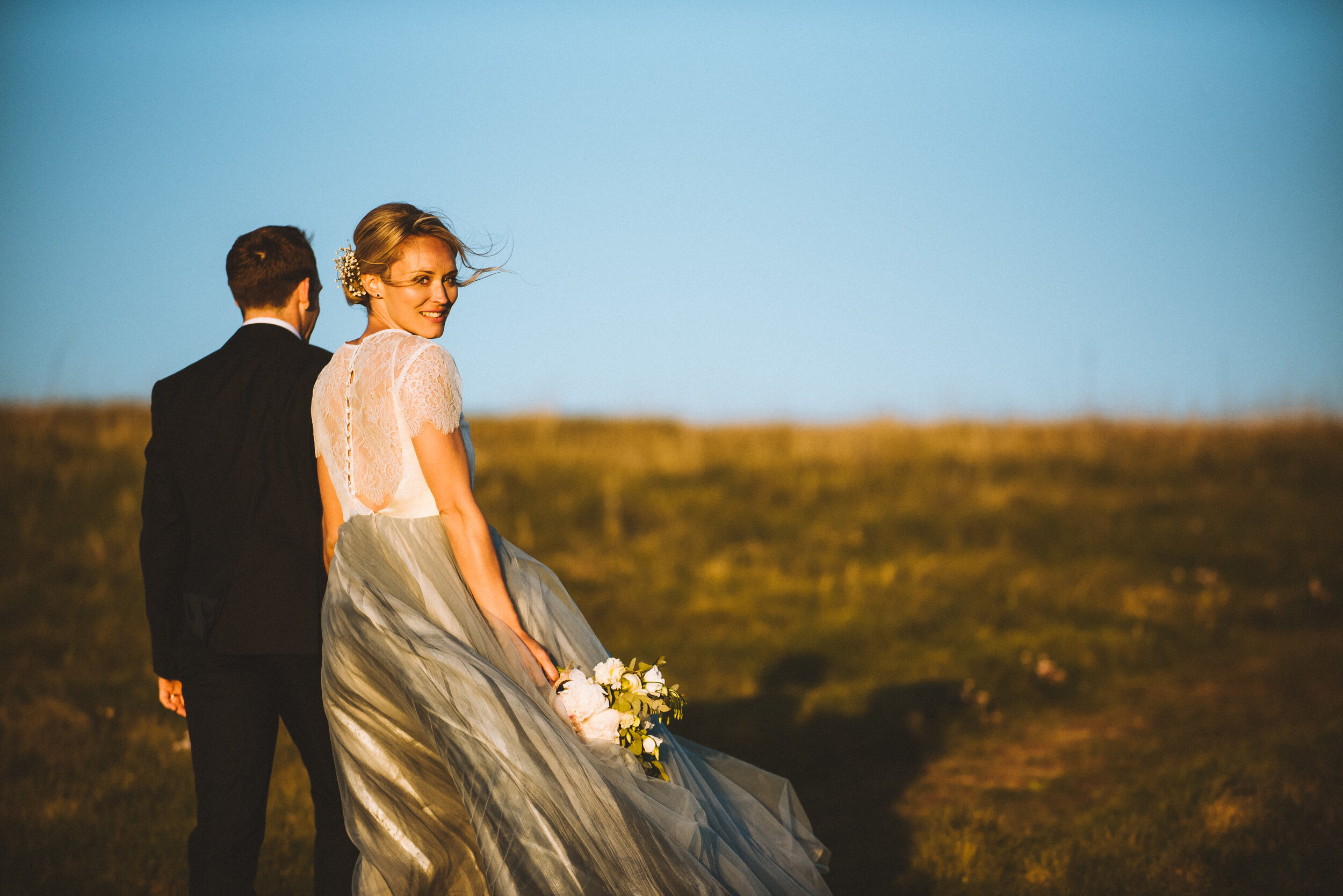 A bride and groom in wedding attire standing outdoors on a grassy hill at sunset, with the bride smiling and holding a bouquet of flowers.