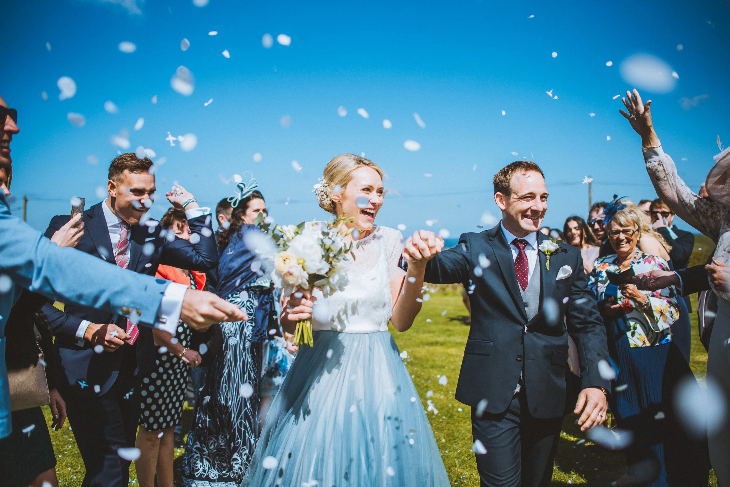 Bride and groom walking outdoors under blue sky, celebrating with friends and family as confetti falls.