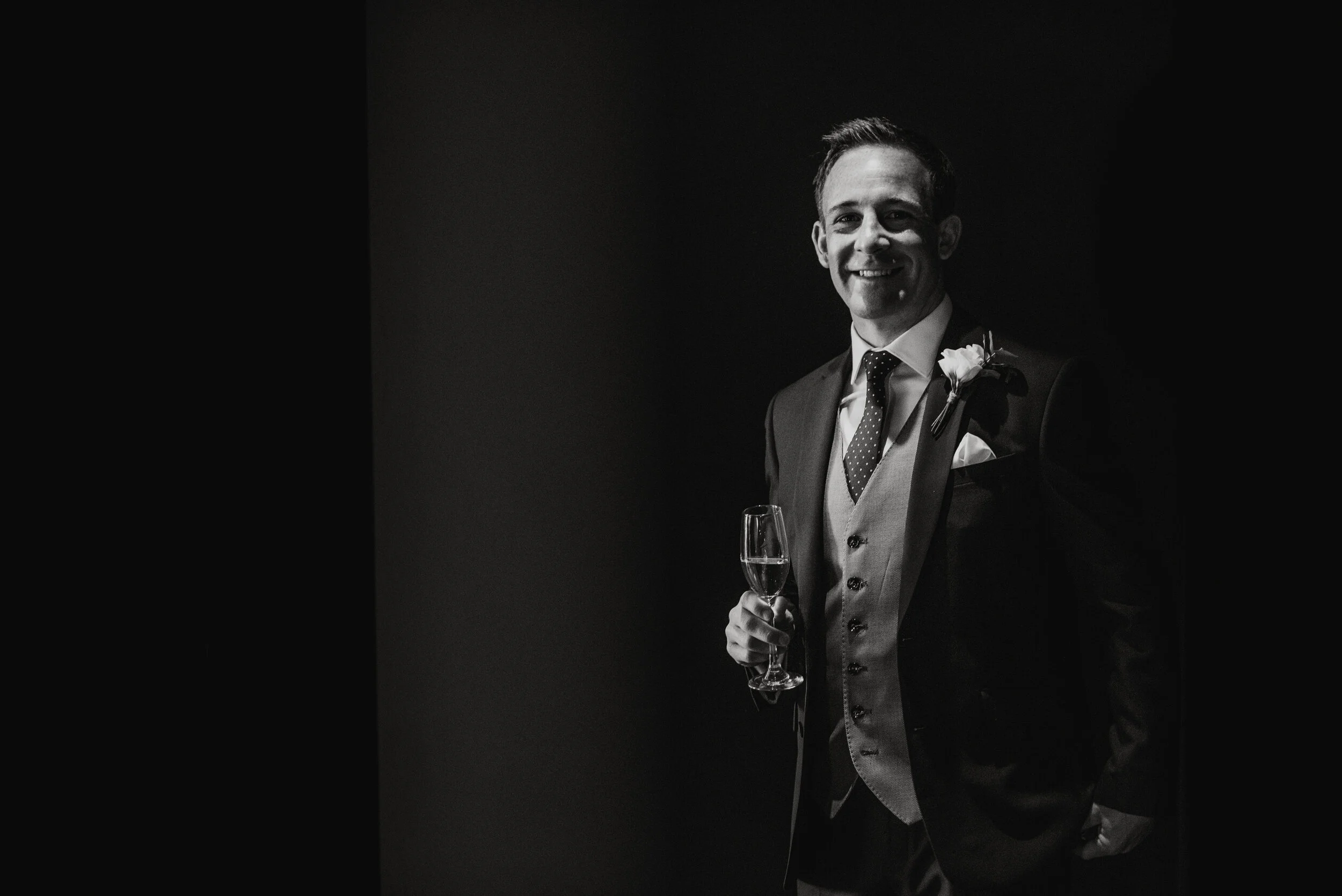 A smiling man in a suit holding a glass of champagne, standing against a dark background.
