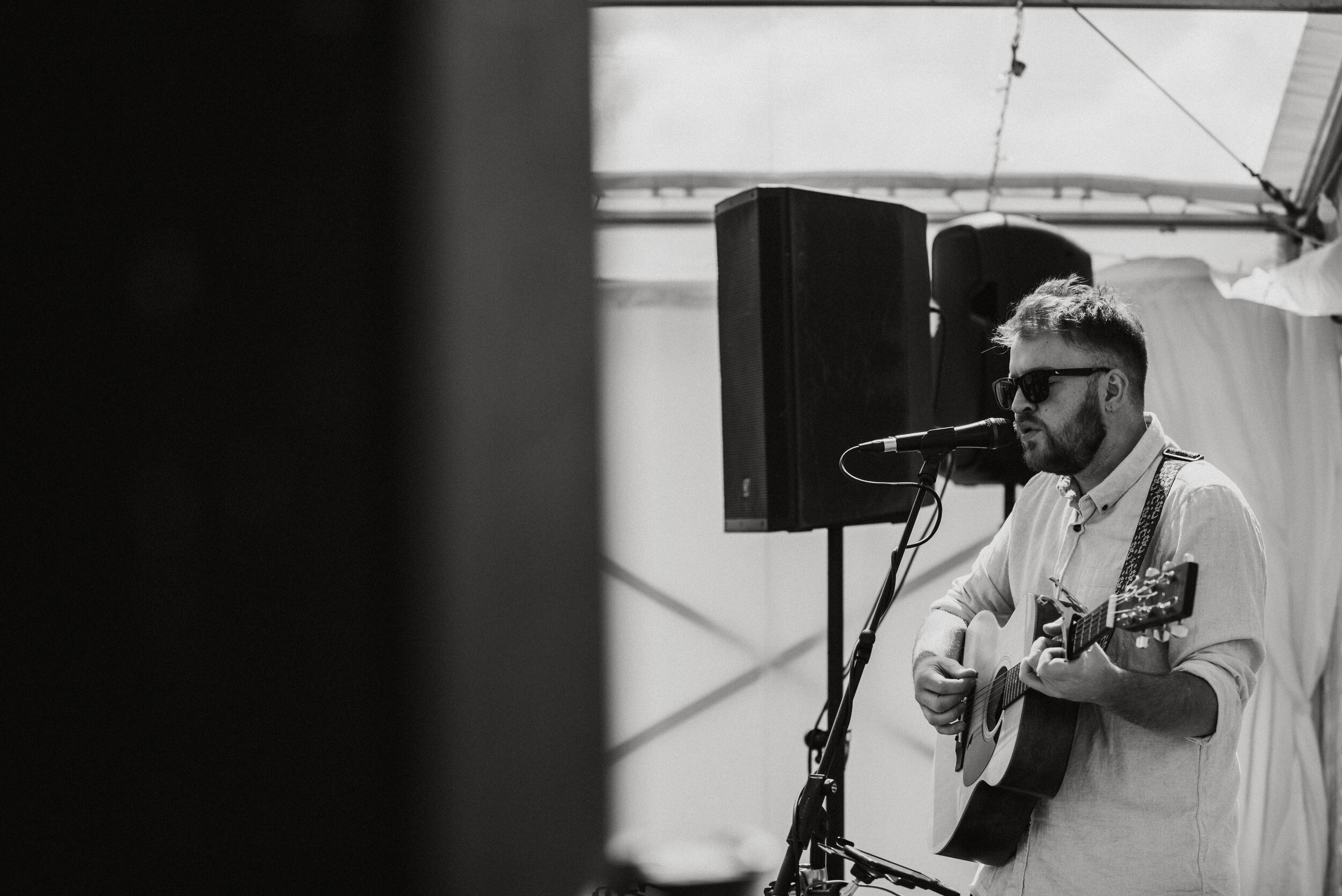 A man with sunglasses singing into a microphone while playing an acoustic guitar, standing near a speaker at an outdoor event.