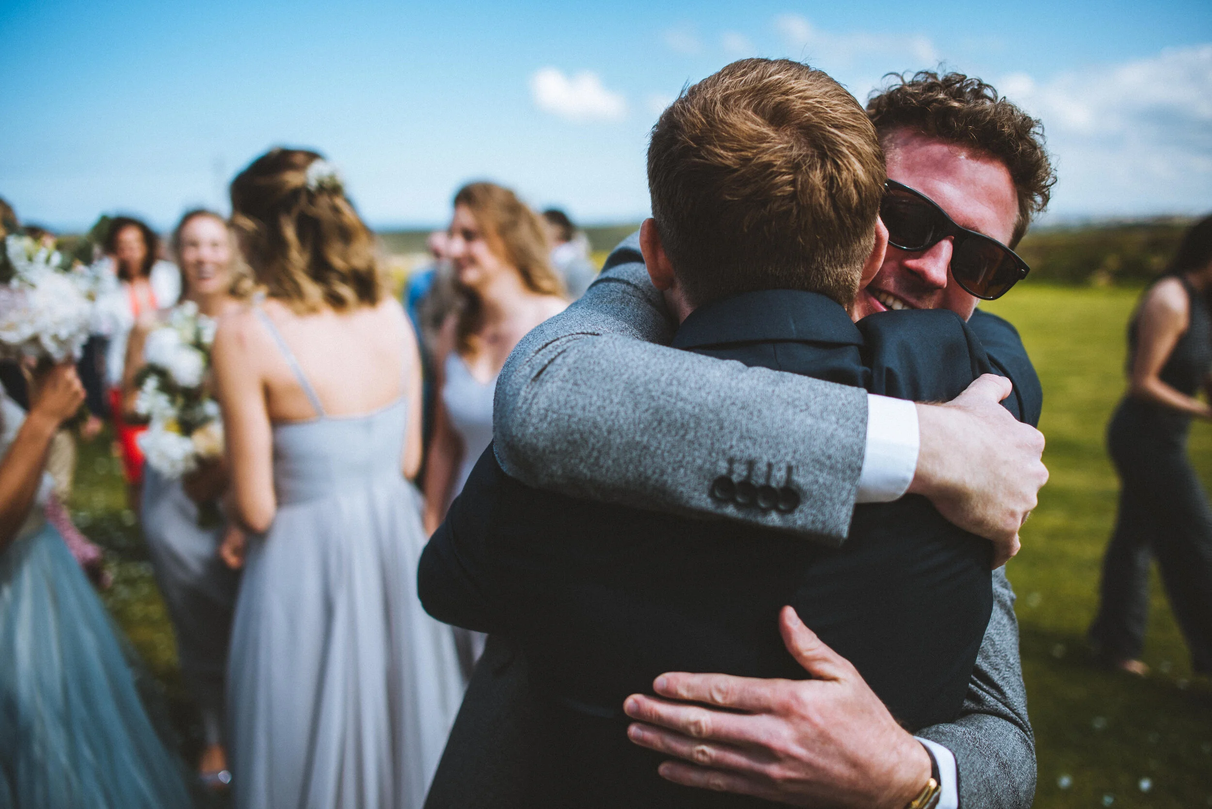 Two men hugging and smiling at an outdoor wedding celebration, with women in dresses and flowers visible in the background.