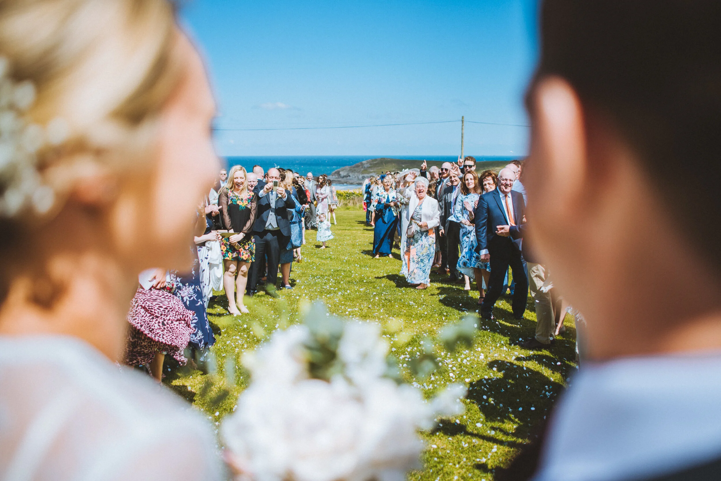 Blurred image of a bride and groom facing each other at an outdoor wedding ceremony, with guests gathered in the background on a sunny day near the coast.
