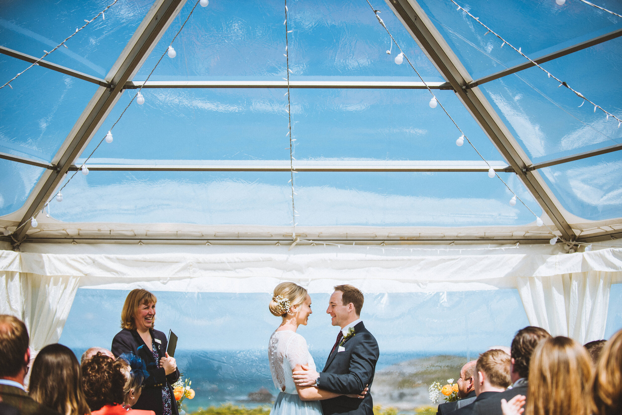 A bride and groom standing close, smiling at each other during their outdoor wedding ceremony under a transparent tent with string lights, with guests seated around them and a scenic view of the coastline in the background.