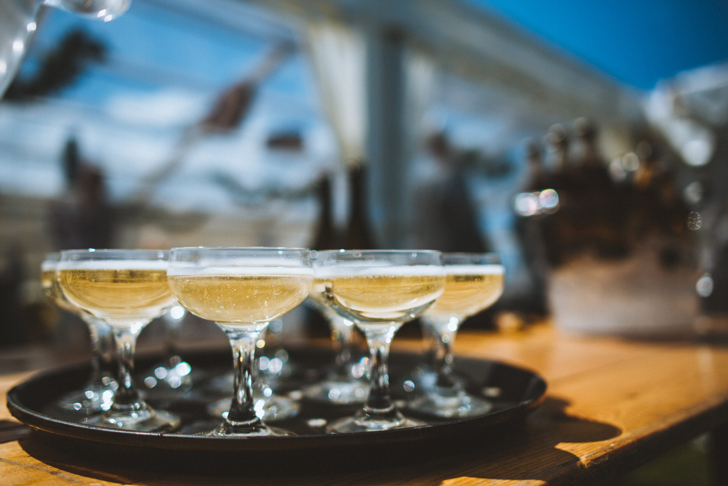 Five coupe glasses filled with champagne on a black tray, on a wooden surface, with a blurred background.