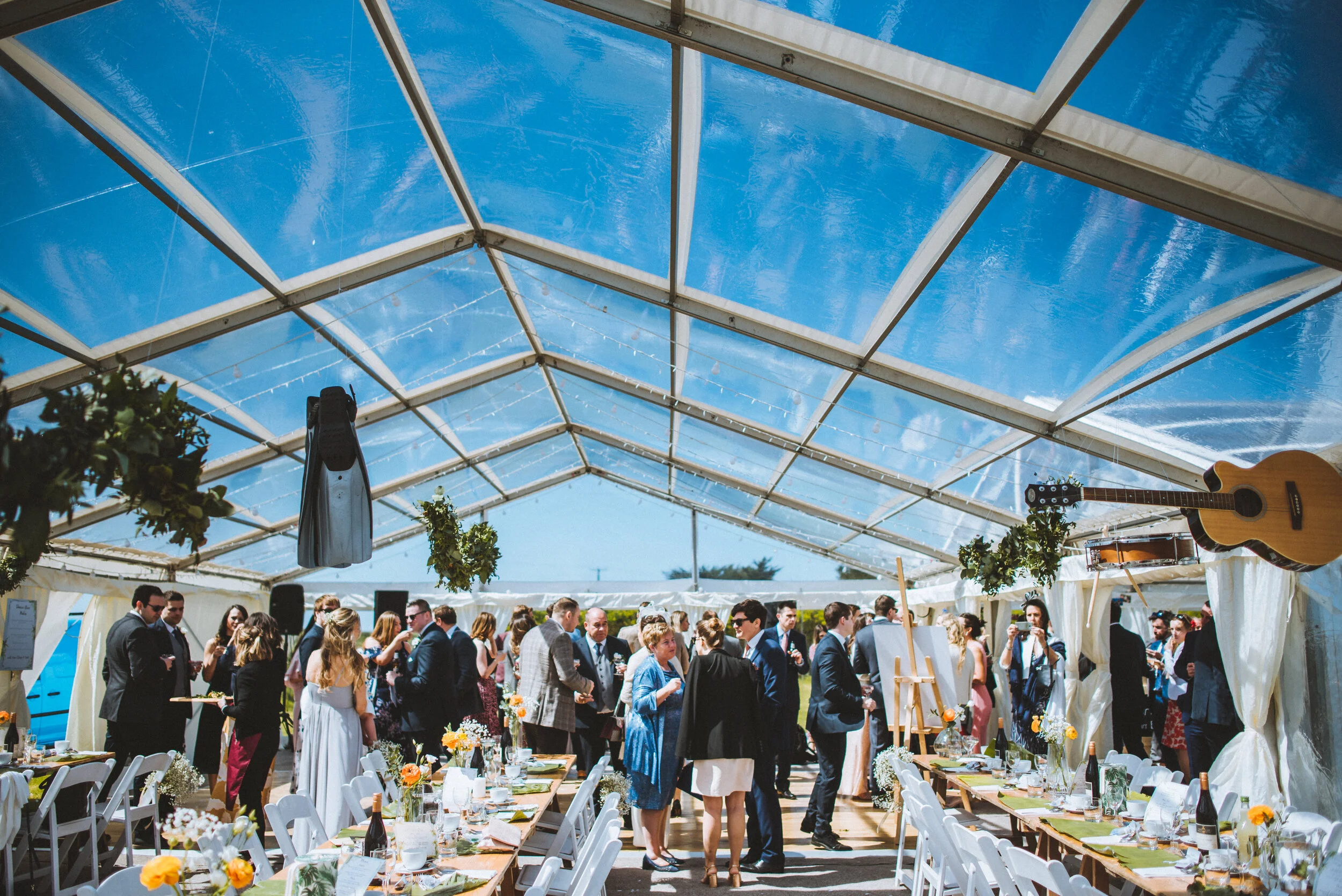Guests at a wedding reception inside a clear tent with a blue sky overhead, decorated with flowers and acoustic guitars hanging from the ceiling, and tables set for dining.