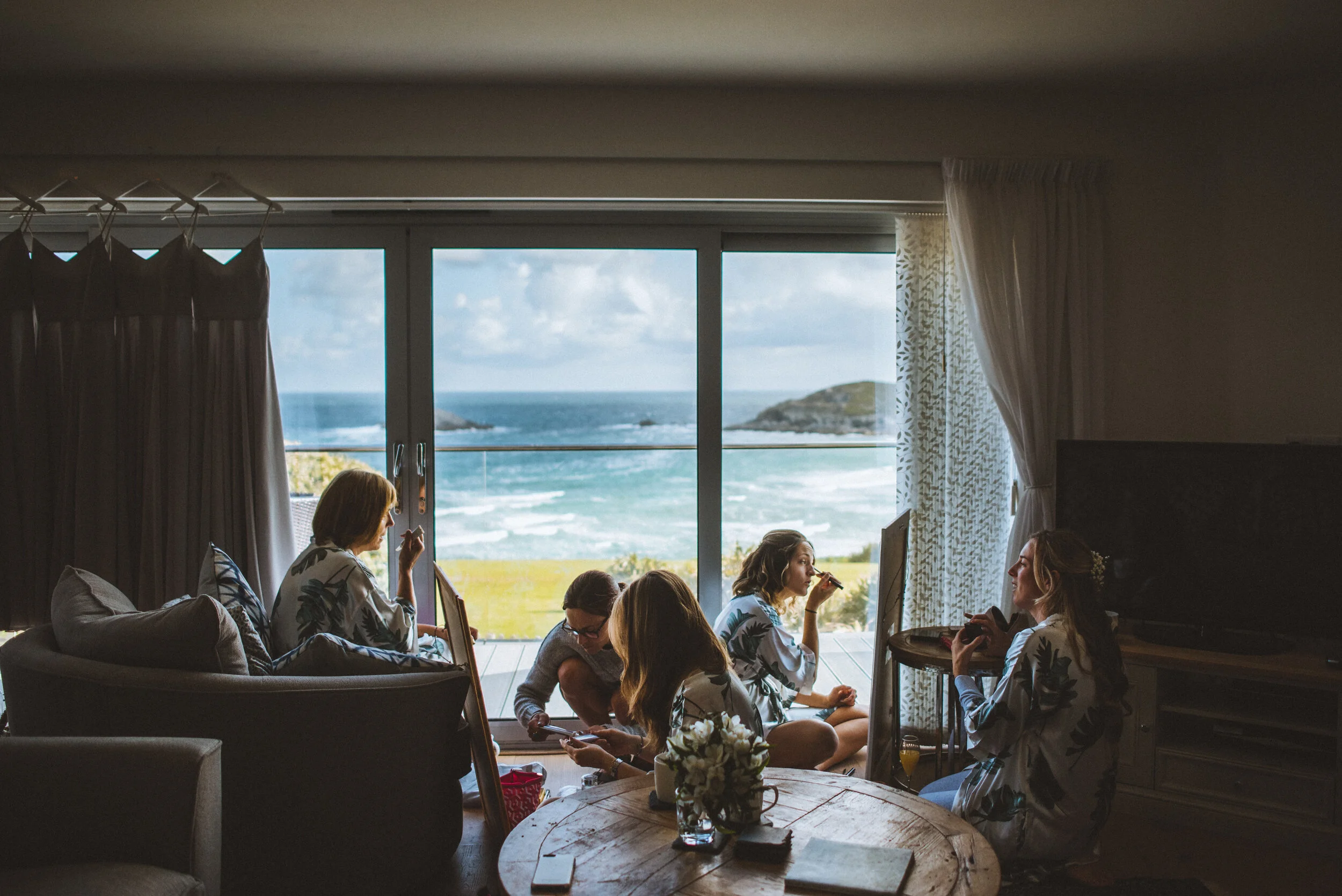 Group of women in robes enjoying a coffee break in a living room with large windows overlooking the ocean and beach.