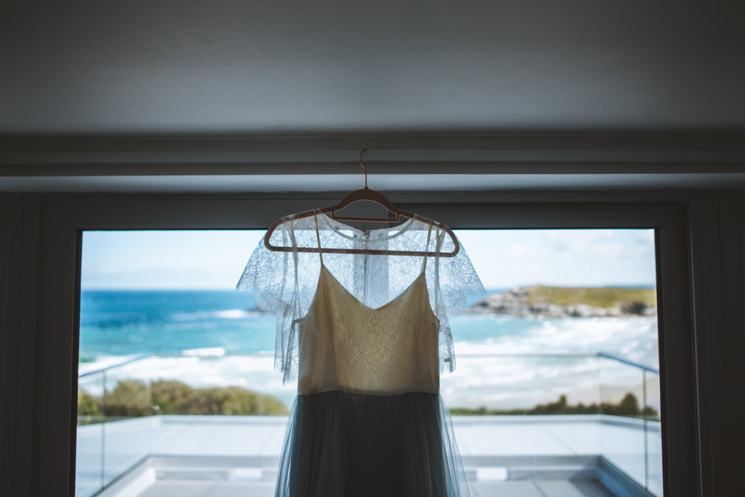 A wedding dress hanging in front of a window overlooking the ocean with a rocky shore and blue sky.