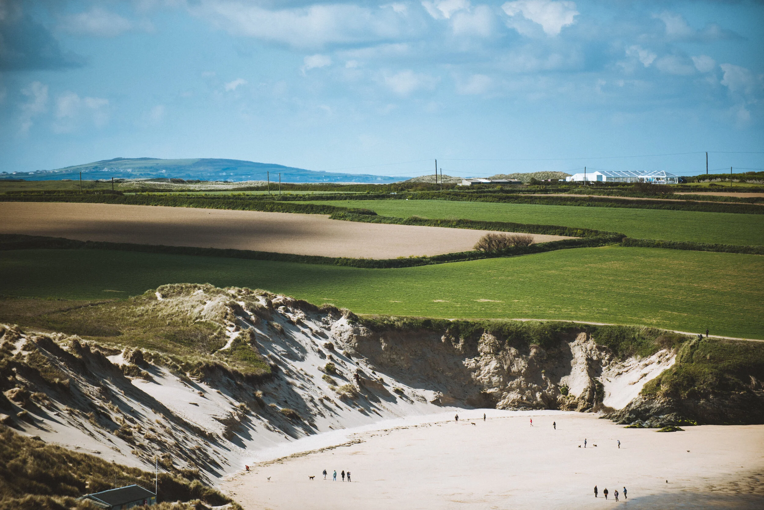 Coastal landscape with green fields, sandy cliffs, and a sandy beach with people and dogs.
