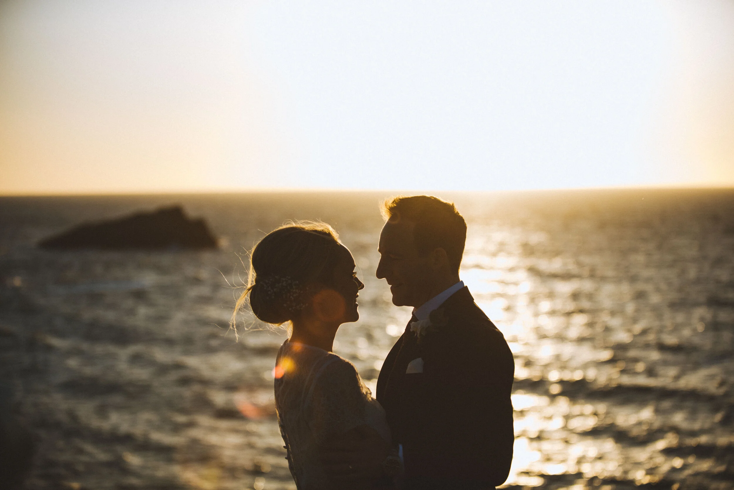 Silhouetted couple at sunset on the beach, facing each other, with ocean and a distant rock formation in the background.