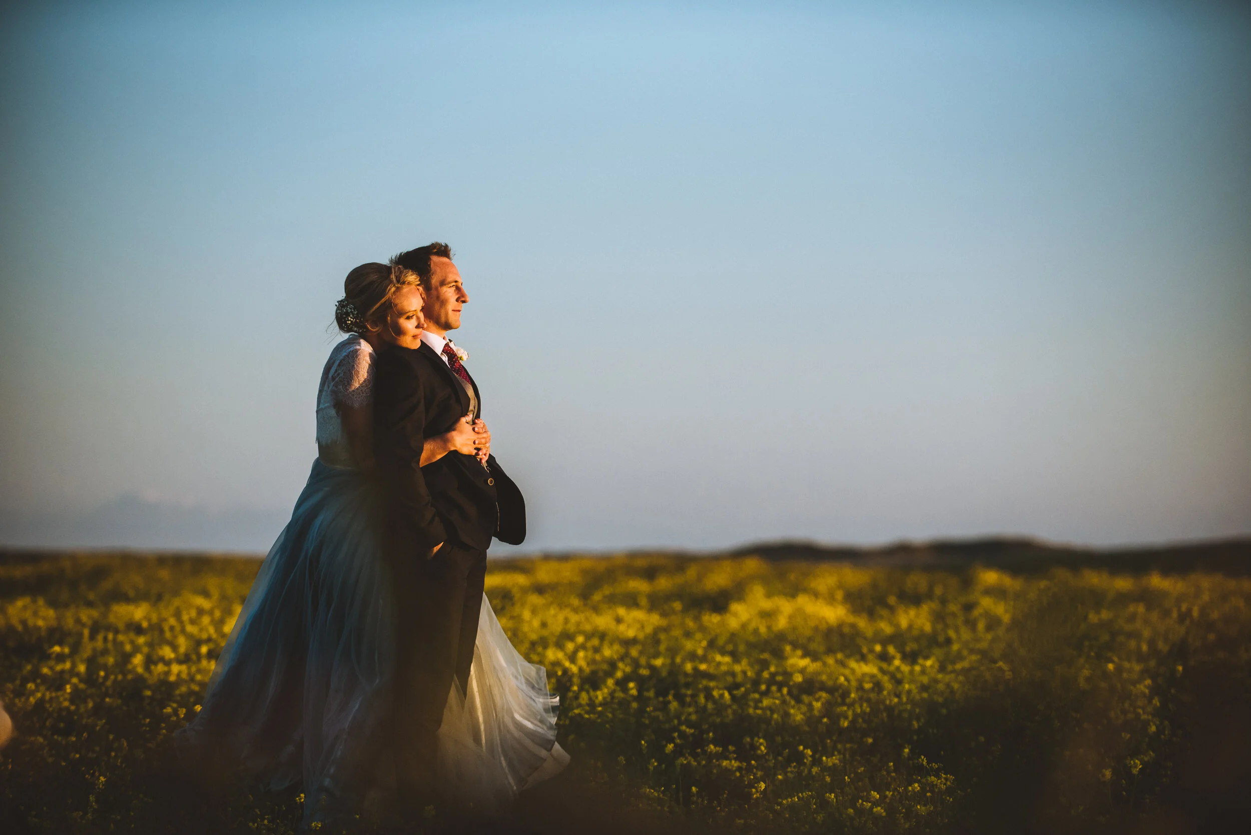 A newlywed couple standing closely in a field of yellow flowers at sunset, with the bride leaning against the groom.