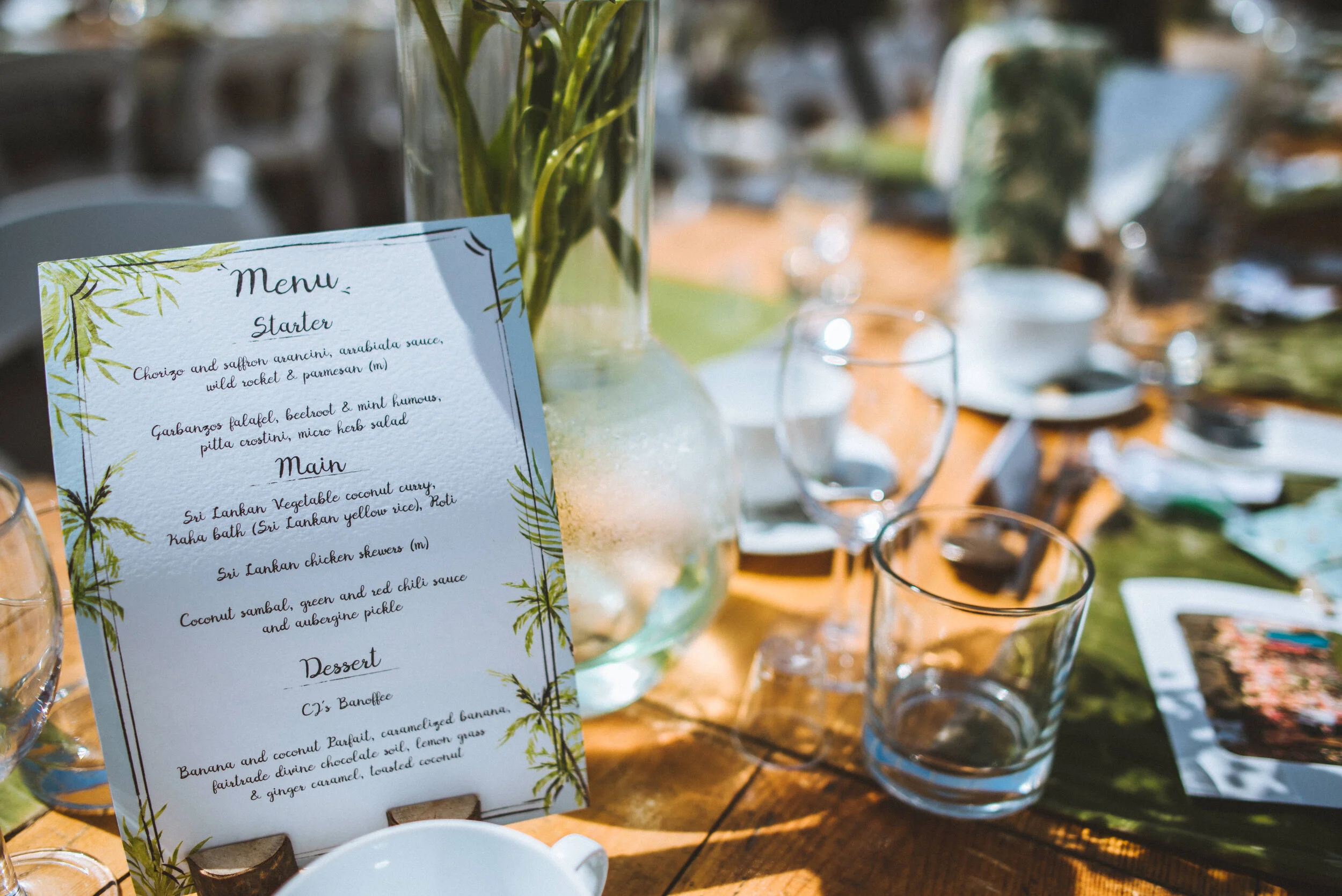 A table with a menu card, clear glasses, a vase with greenery, and a sunlight-lit outdoor setting.