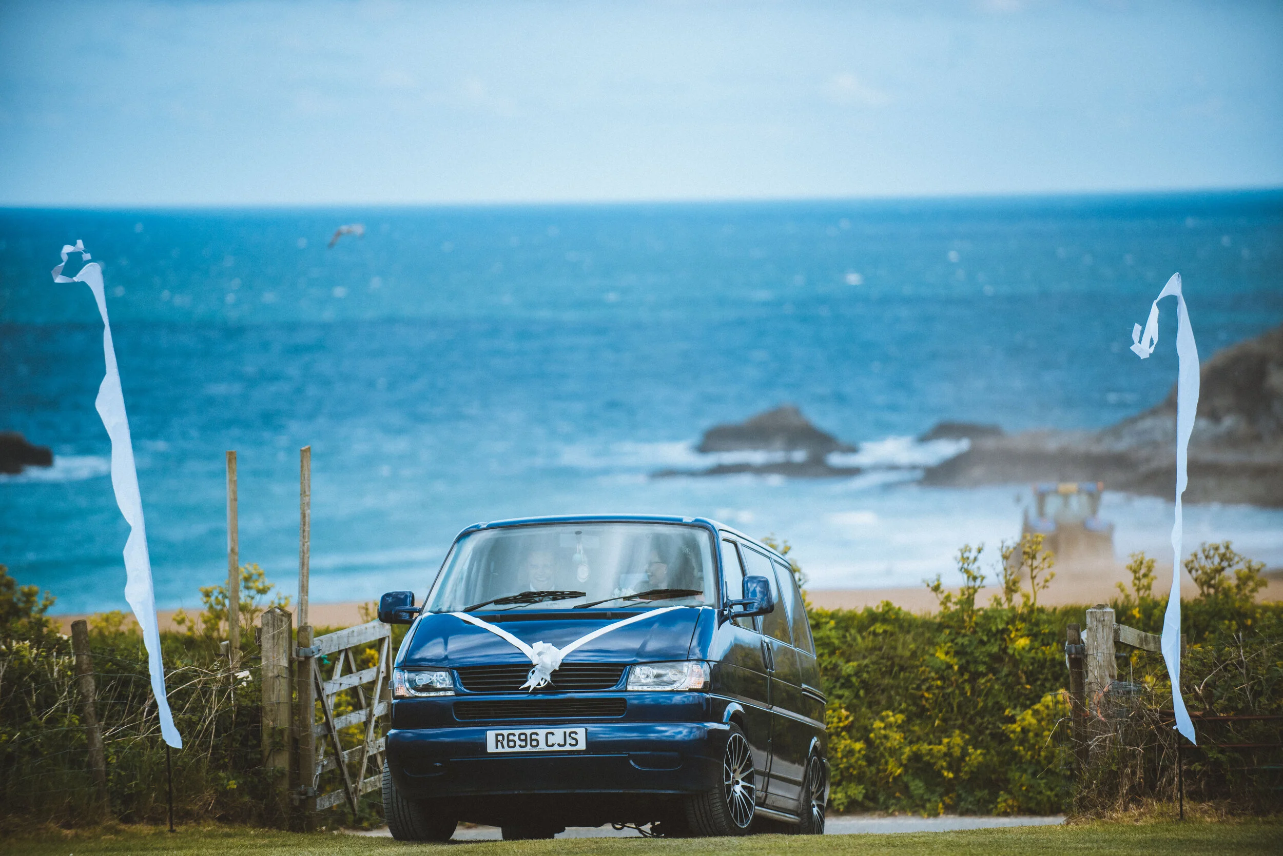 Black van decorated with white ribbons in front of a coastal view with the ocean, rocky formations, and overcast sky.
