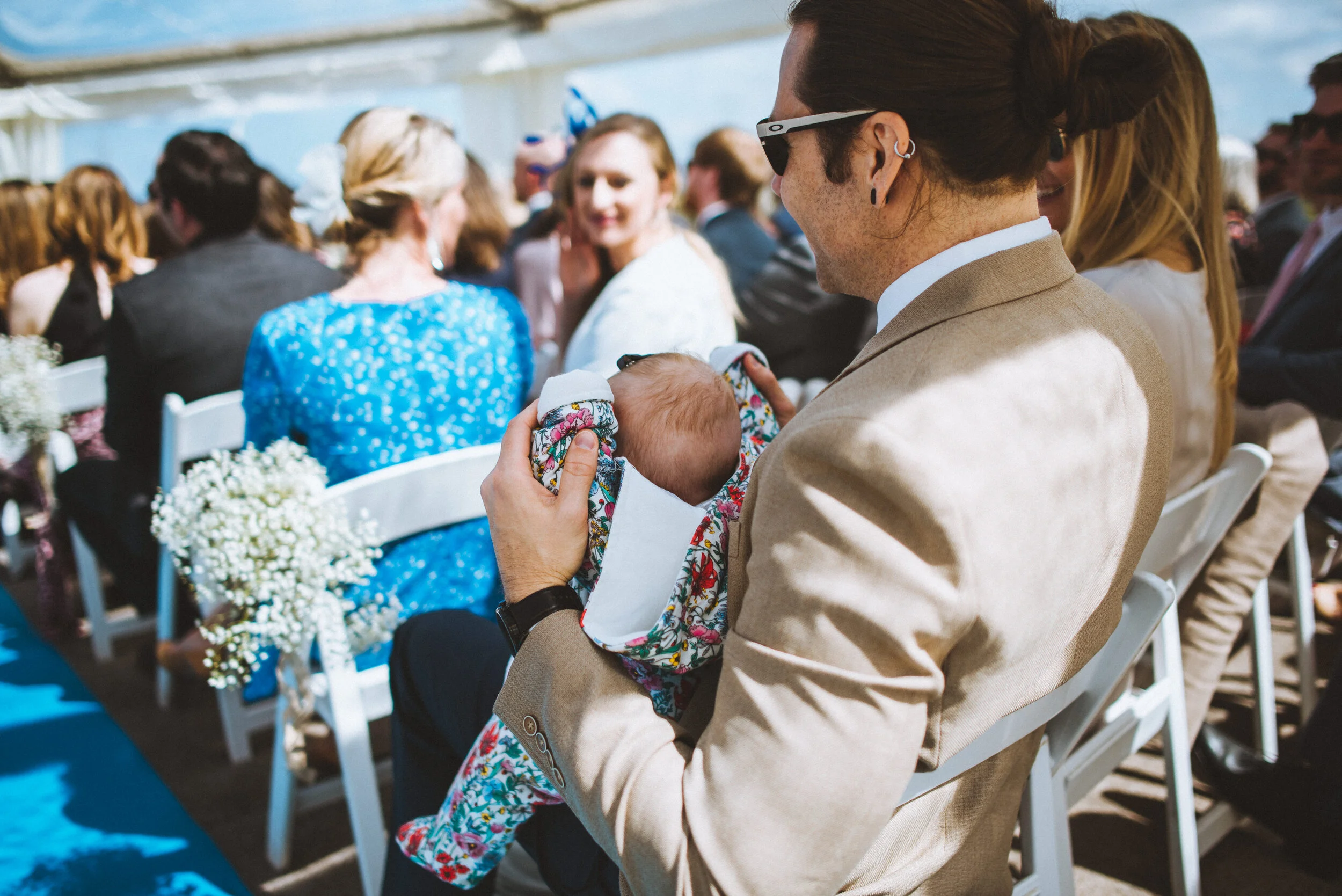A man in a beige suit holds a baby wrapped in a colorful floral blanket at an outdoor wedding ceremony, with guests seated in white chairs around him.