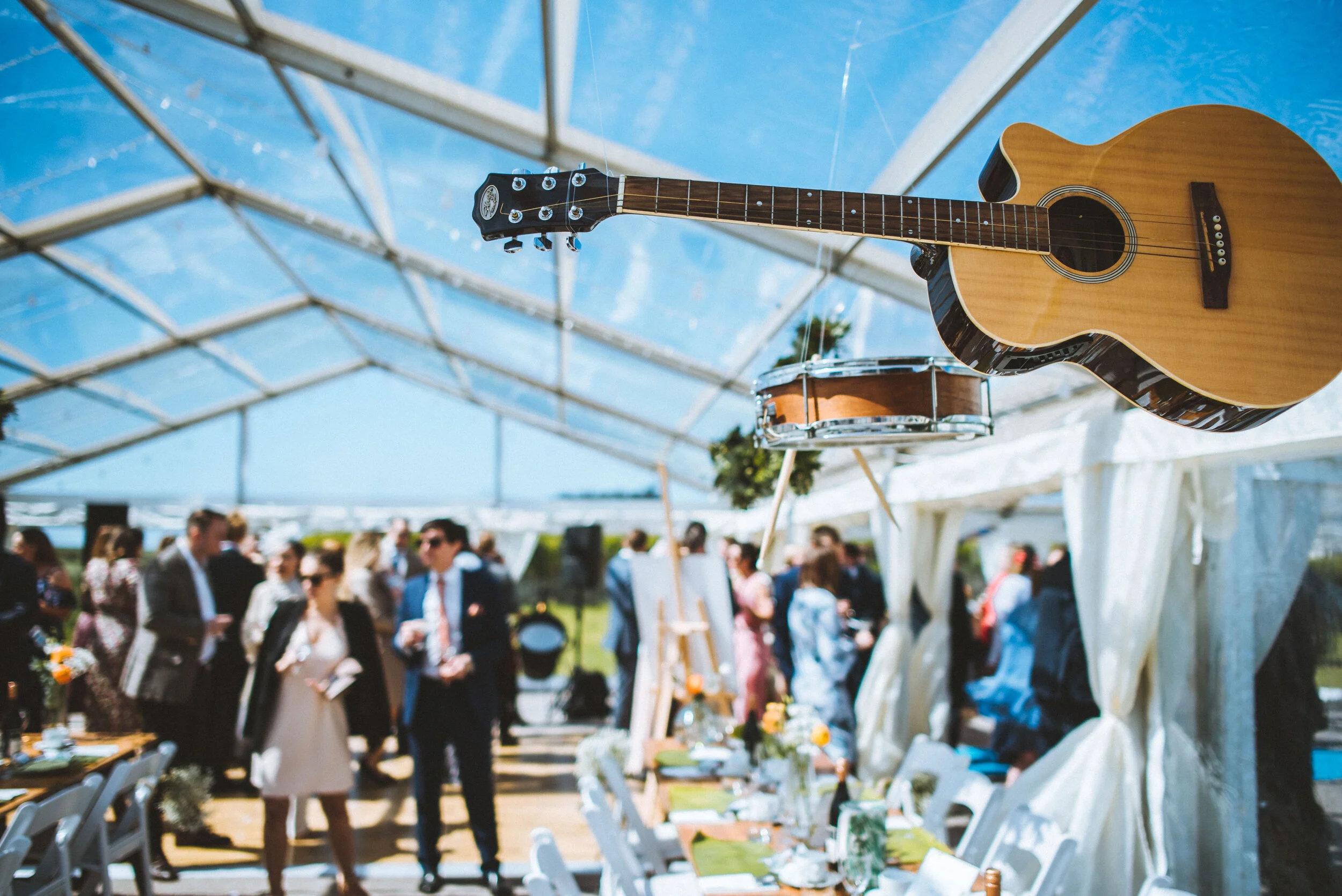 A guitar and a tambourine hanging inside a bright, transparent tent at a lively outdoor event with many people socializing and dressed in formal attire.