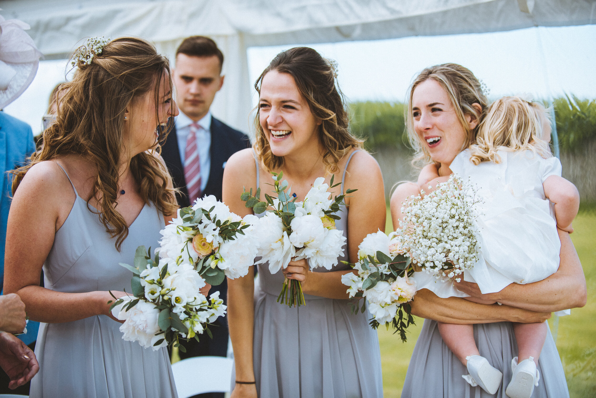 People dressed in formal attire holding floral bouquets at an outdoor wedding celebration.