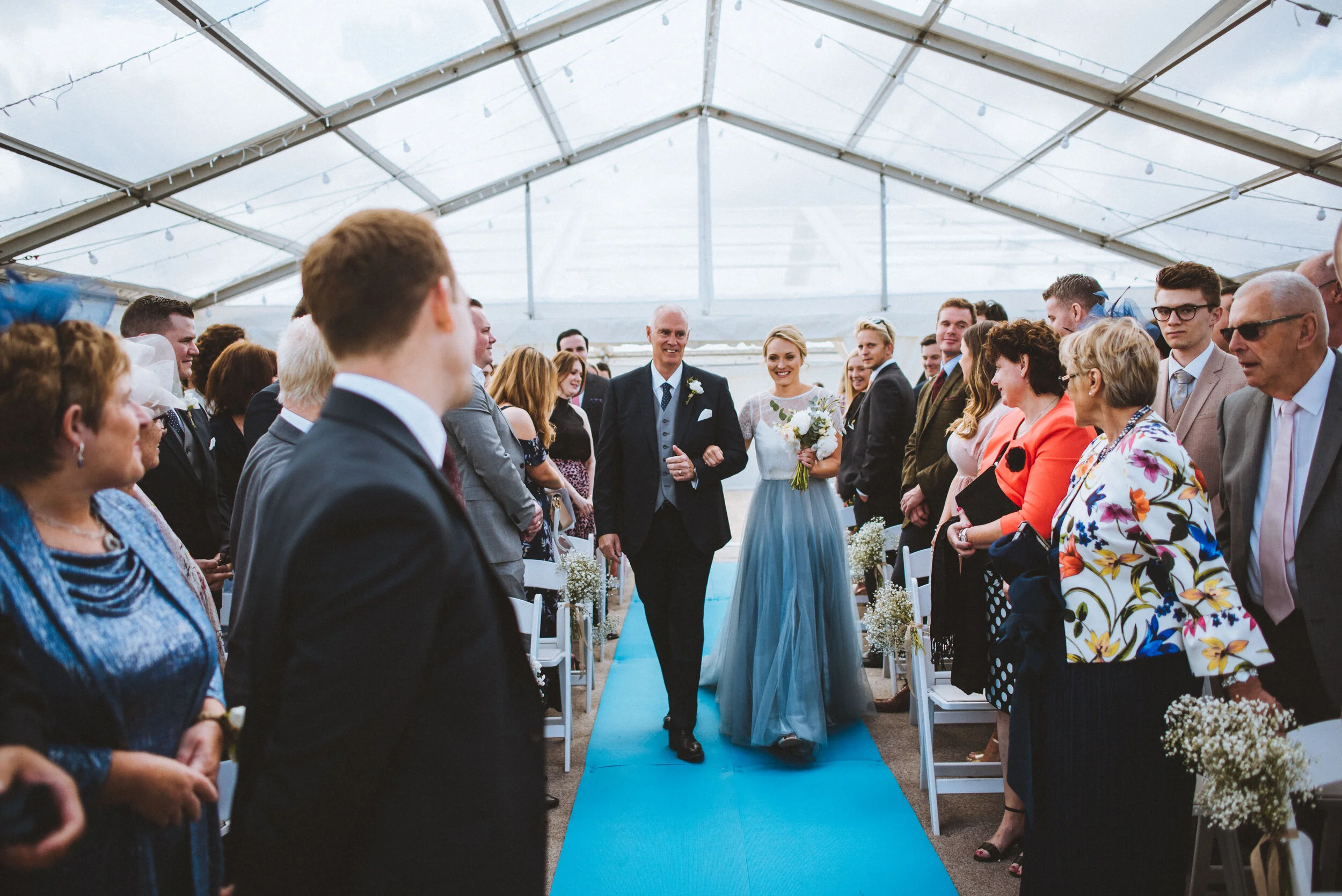 A bride walking down the aisle with her father at a wedding ceremony inside a glass tent.