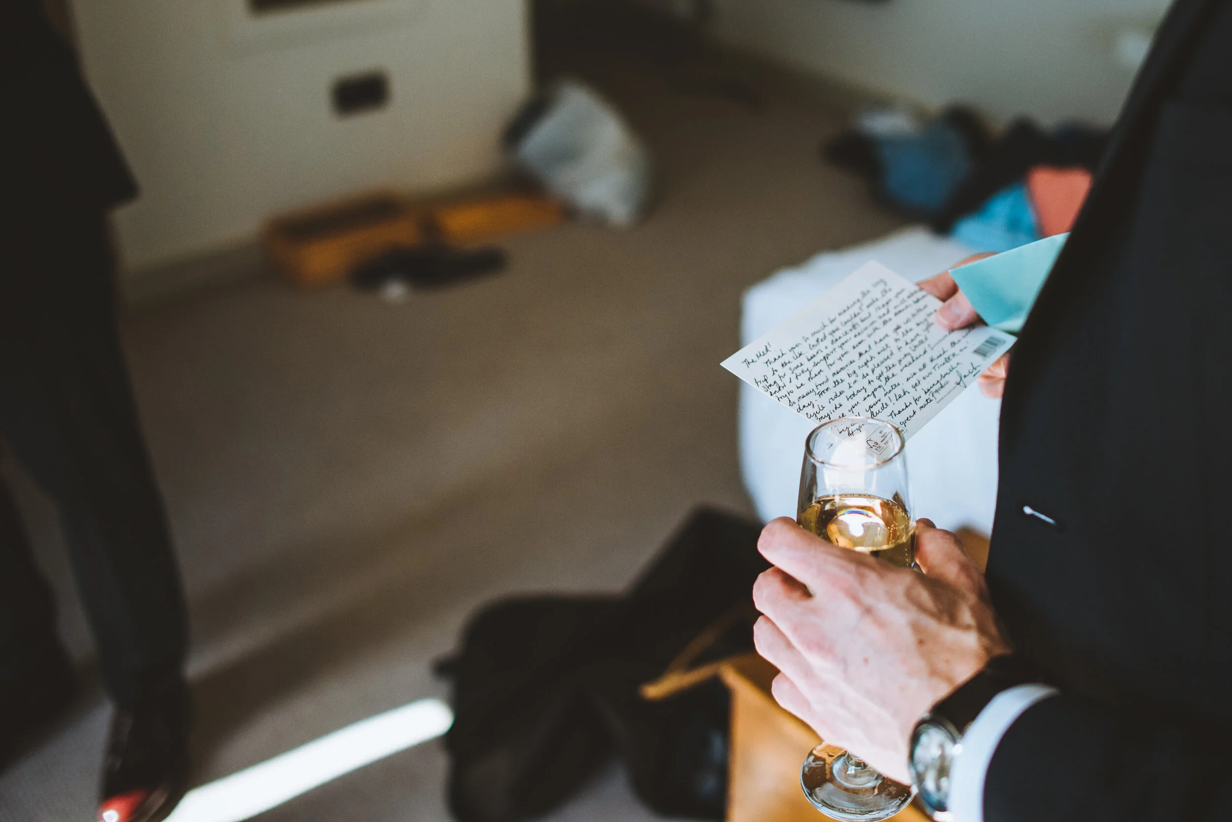 Person holding a glass of champagne and a handwritten letter in a room with a beige carpet and some scattered items on the floor.