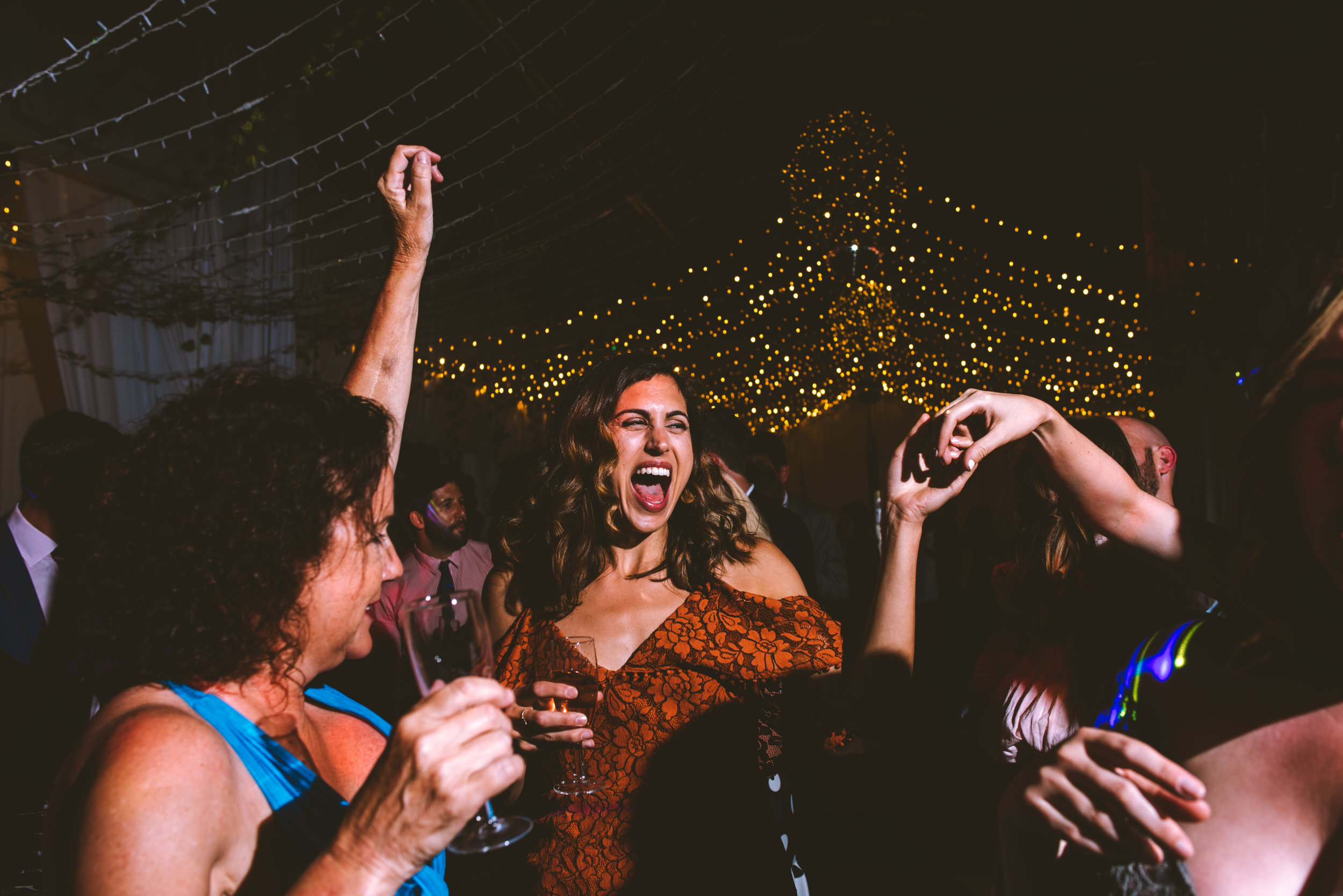 People dancing and celebrating at a party with string lights hanging overhead, a woman in a floral dress singing or shouting, and others holding drinks, in a festive, lively atmosphere.