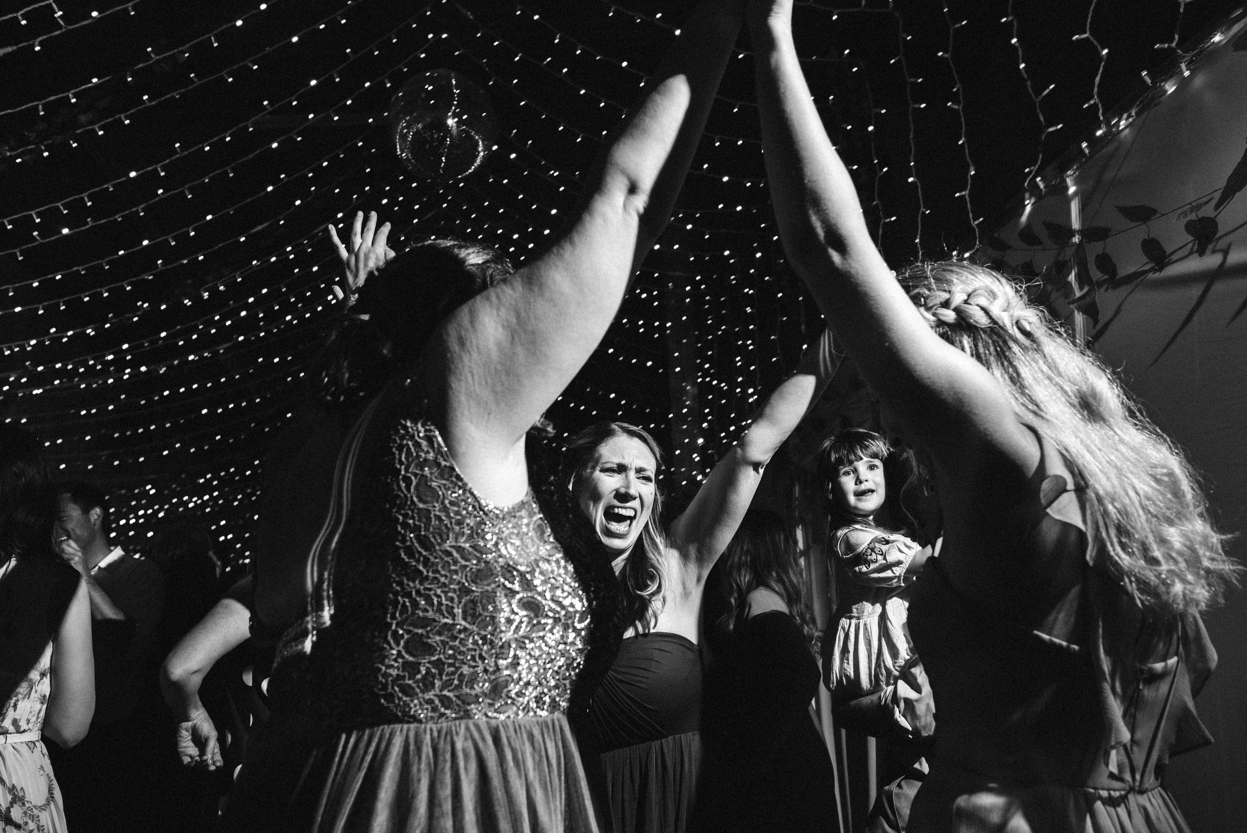 People dancing and celebrating at a party or wedding under string lights, with visible expressions of joy and excitement.