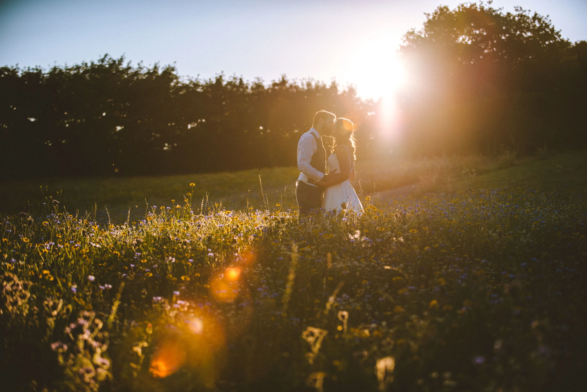 A couple standing close together in a field of flowers during sunset, embracing each other with the sun shining behind them.