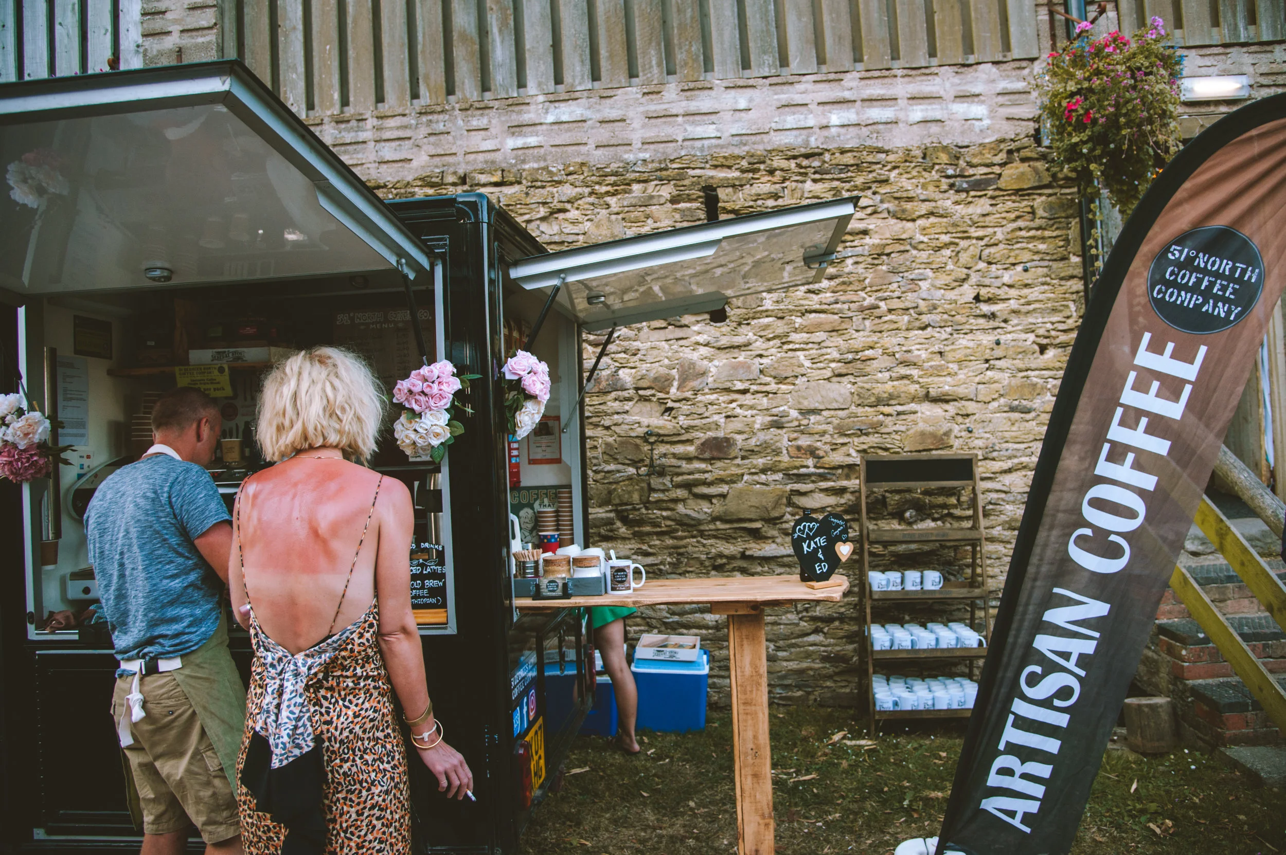 People ordering coffee at an artisan coffee truck outdoors against a brick wall, with a tall flag announcing artisan coffee and a small chalkboard sign with hearts.