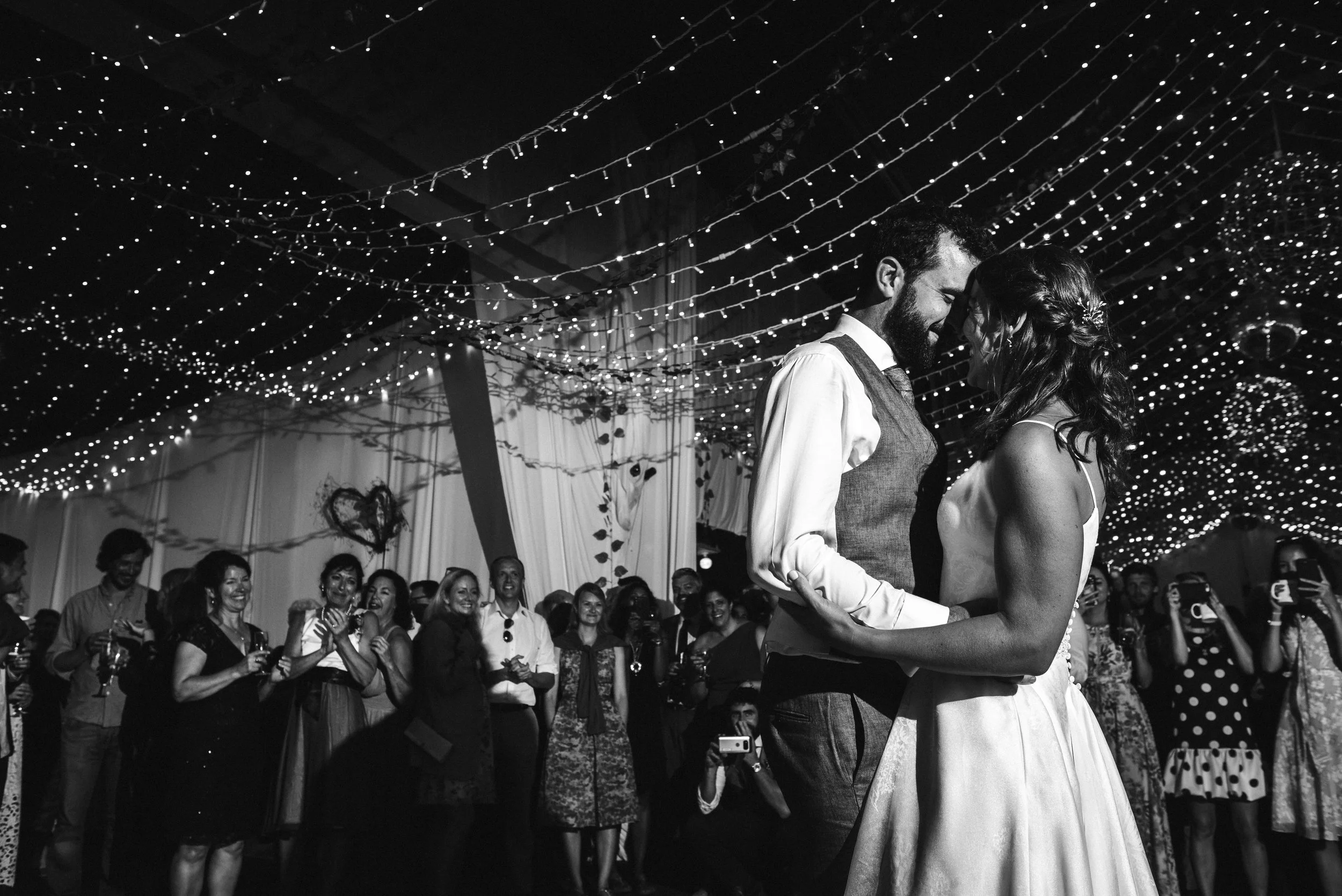 Black and white photo of a couple sharing their first dance at a wedding reception under string lights, with guests watching and taking photos in the background.