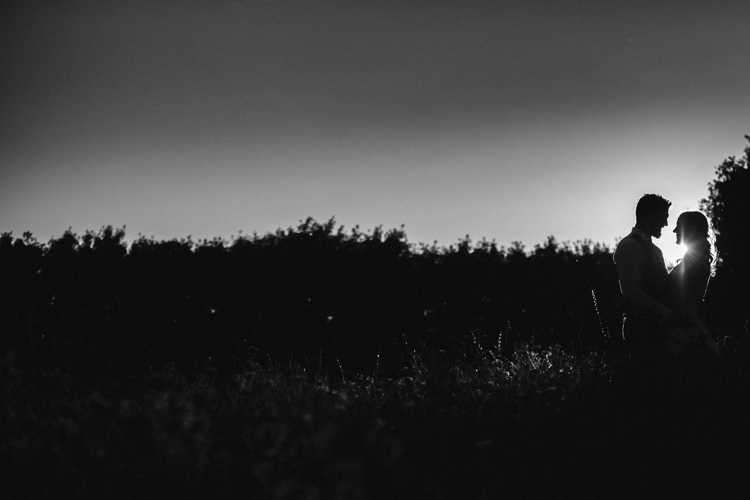 Silhouette of a couple standing close together outdoors at sunset, with the sun behind them creating a backlit effect.
