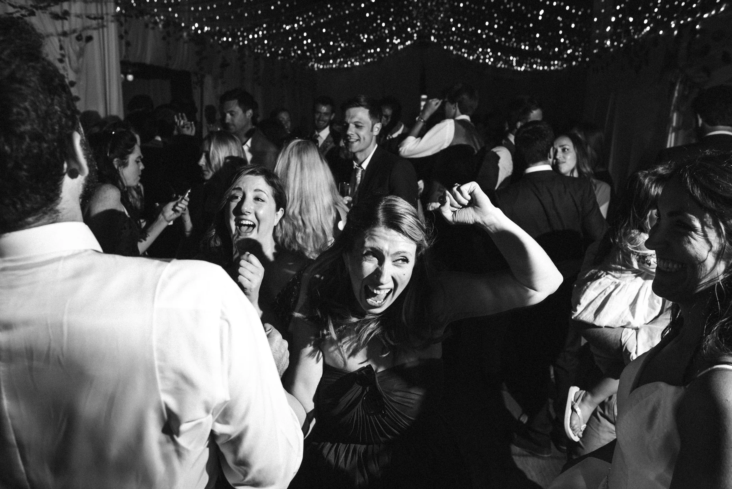Black and white photo of people dancing and celebrating at a party, with hanging string lights overhead.