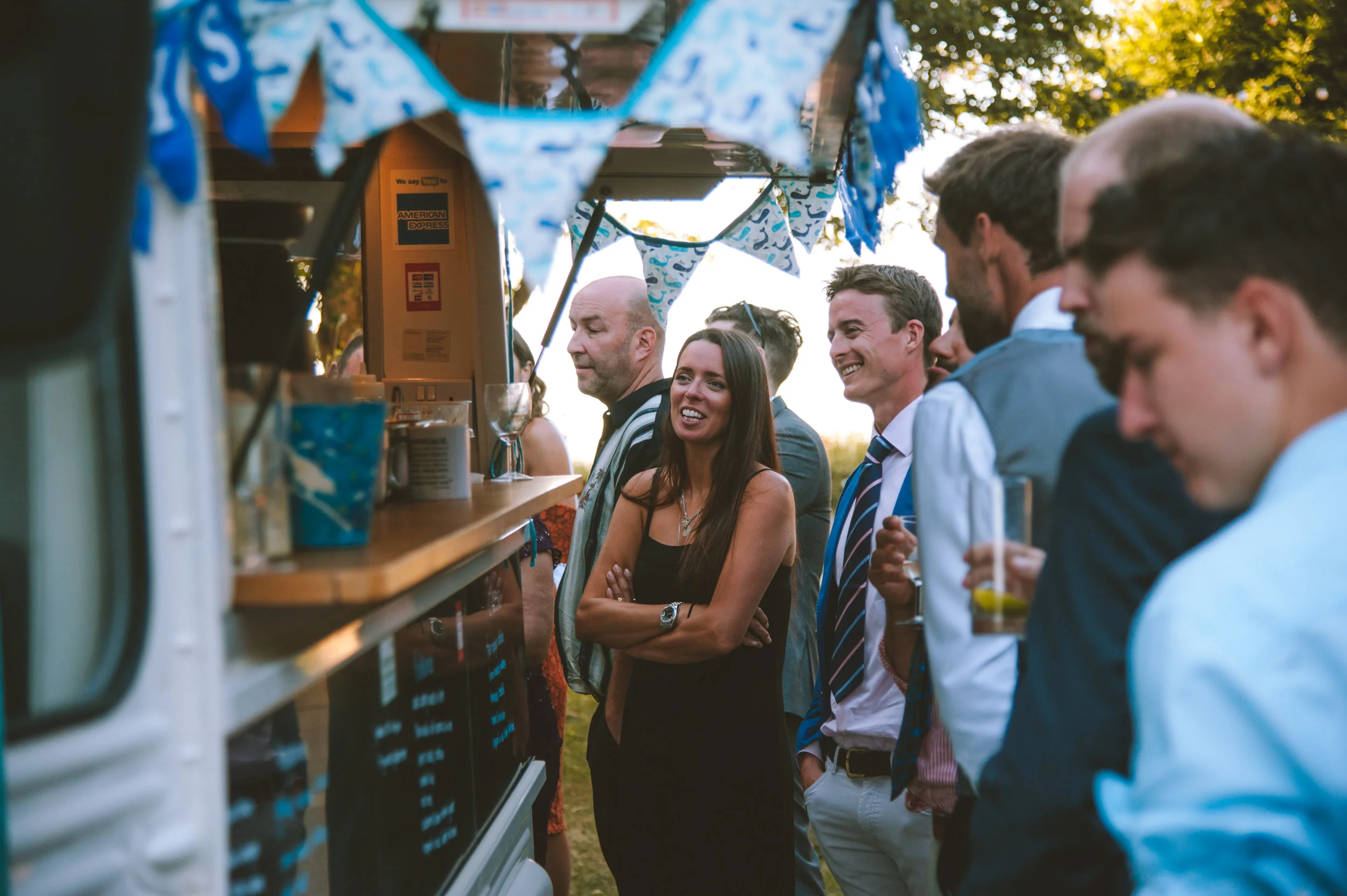 People at an outdoor event standing gathered around a bar cart with American Express branding, decorated with blue and white bunting.