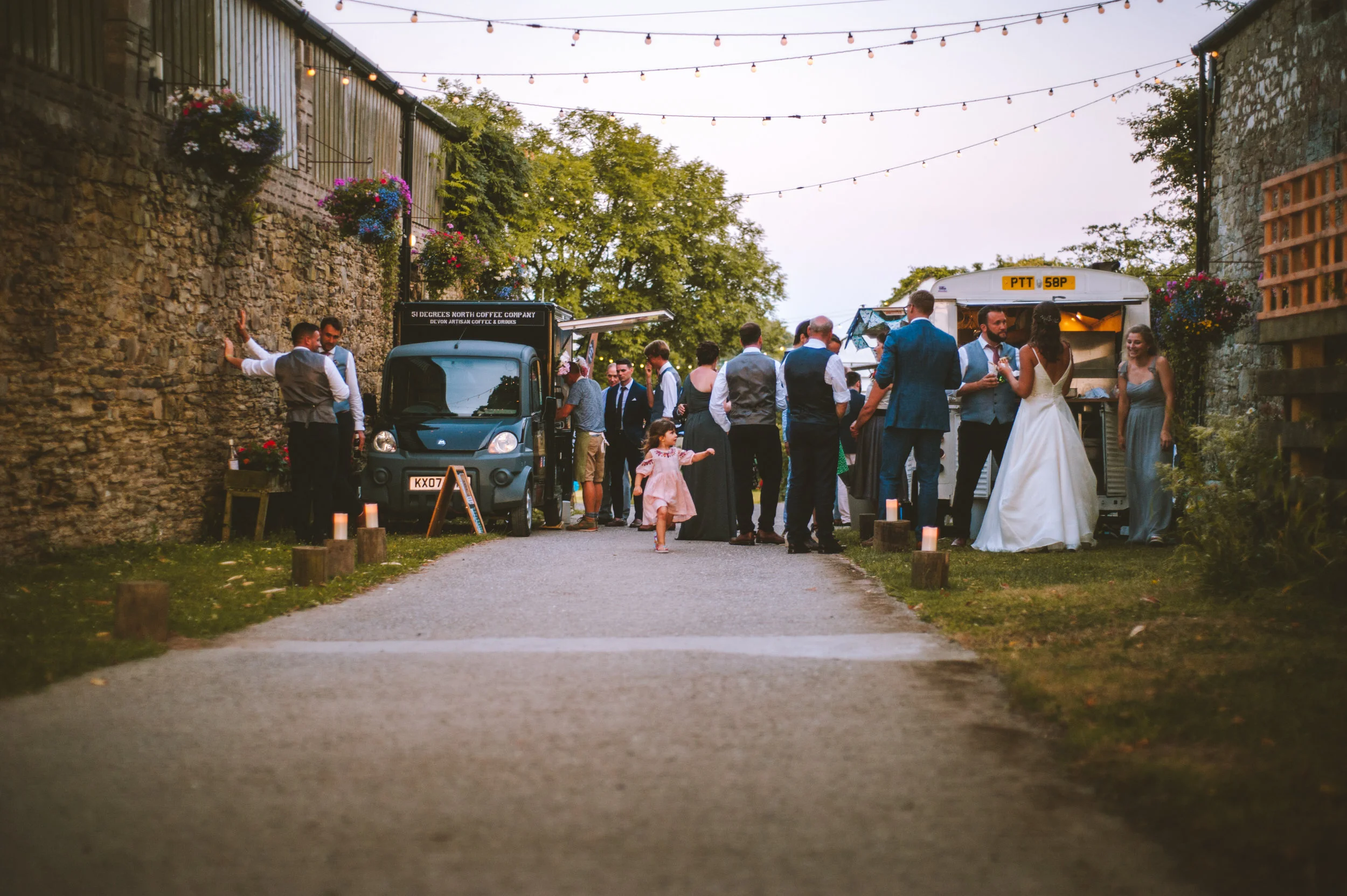 A wedding reception outdoors with guests gathered near a food truck, along a pathway decorated with small candles and string lights hanging above. The background includes stone walls and lush greenery.
