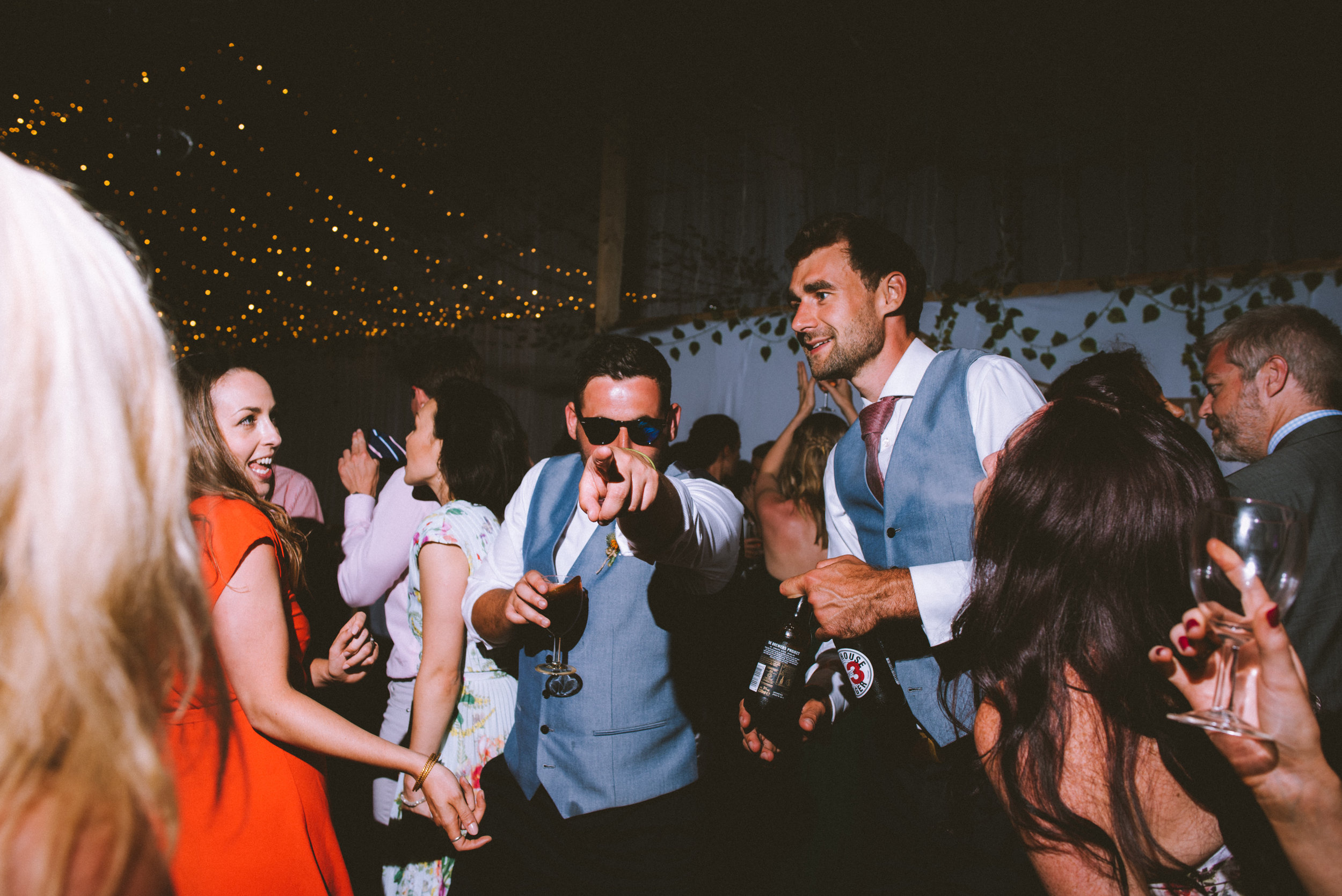 People dancing and socializing at a party or wedding reception, with string lights overhead and a decorated background.