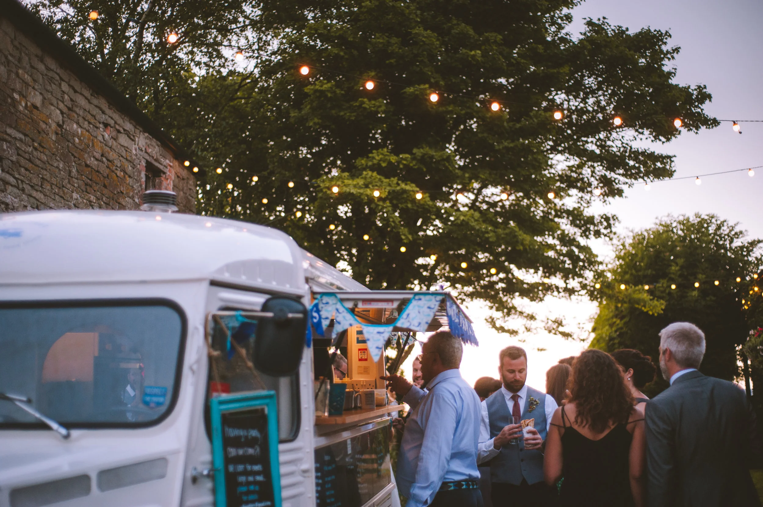 People in formal attire gather around a food truck during an outdoor event at dusk, with string lights overhead and trees in the background.