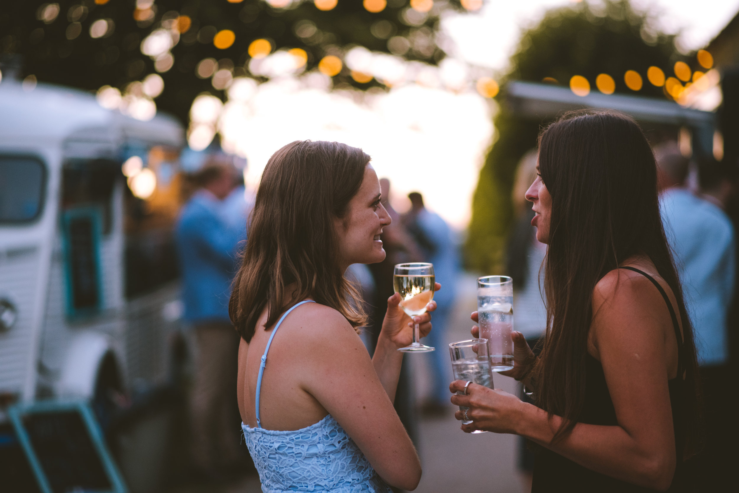 Two women with dark hair talking and smiling at an outdoor event during sunset, holding glasses of wine and water, in a social setting with blurred people and string lights in the background.