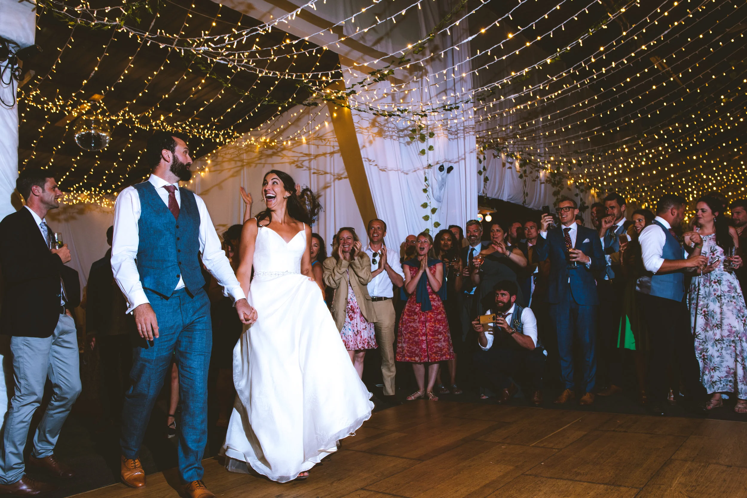 A wedding reception with a bride and groom holding hands and dancing under string lights and hanging decorations, while guests cheer and watch in the background.