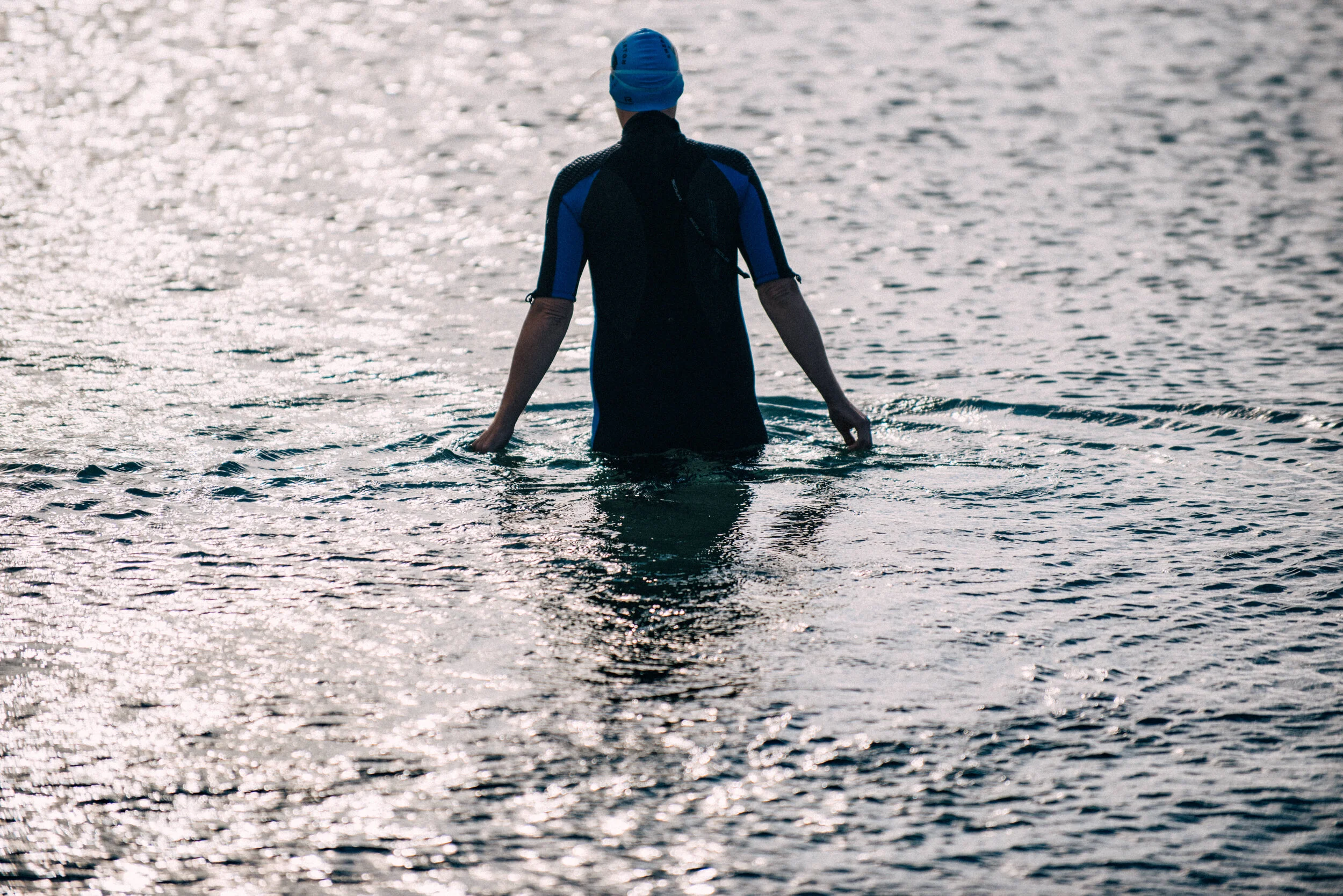 A person wearing a blue swim cap and a wetsuit standing in calm water, facing away from the camera.