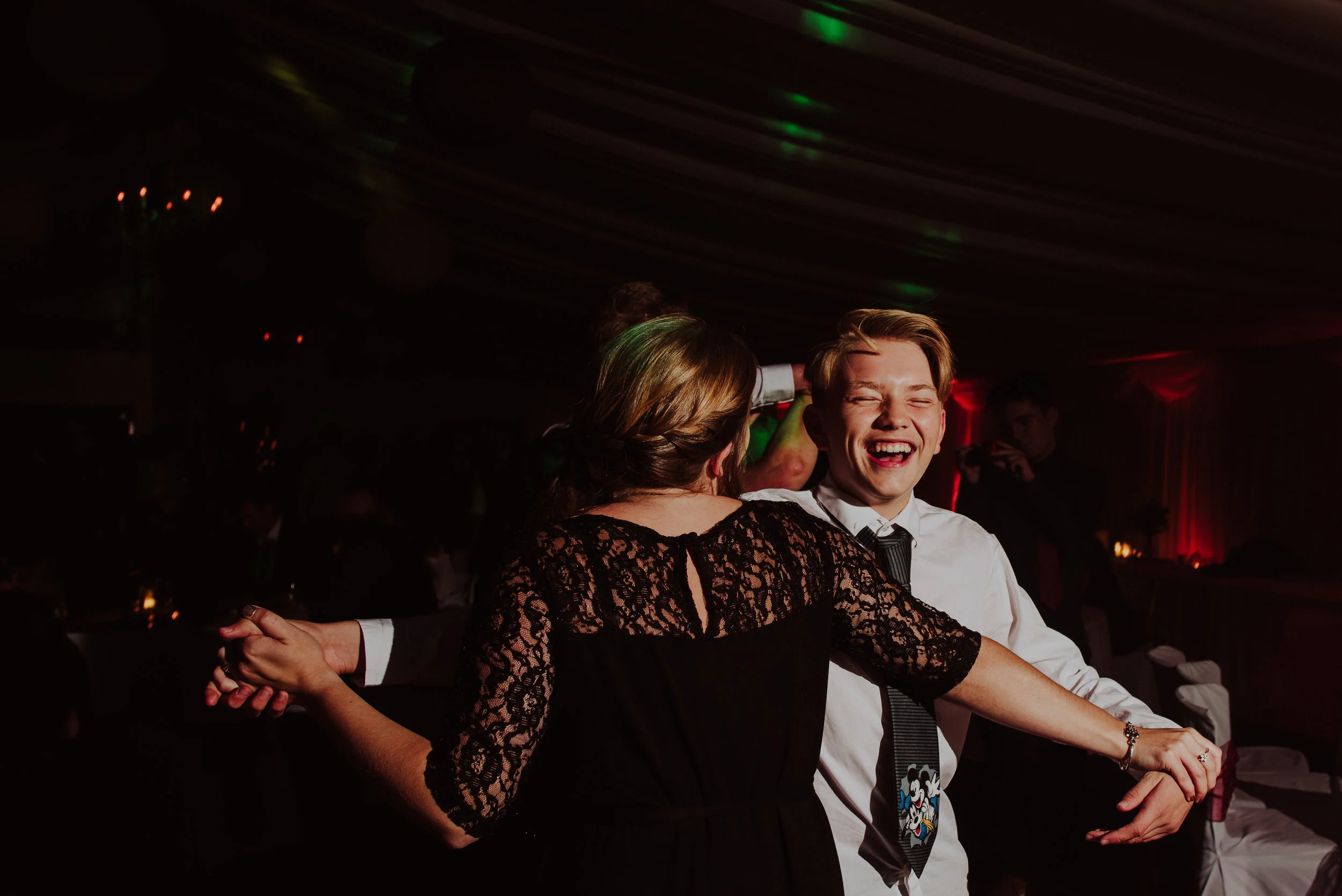 A young man and woman dancing at a celebration or party, with the man smiling and the woman facing away, in a dimly lit room with colorful lights.