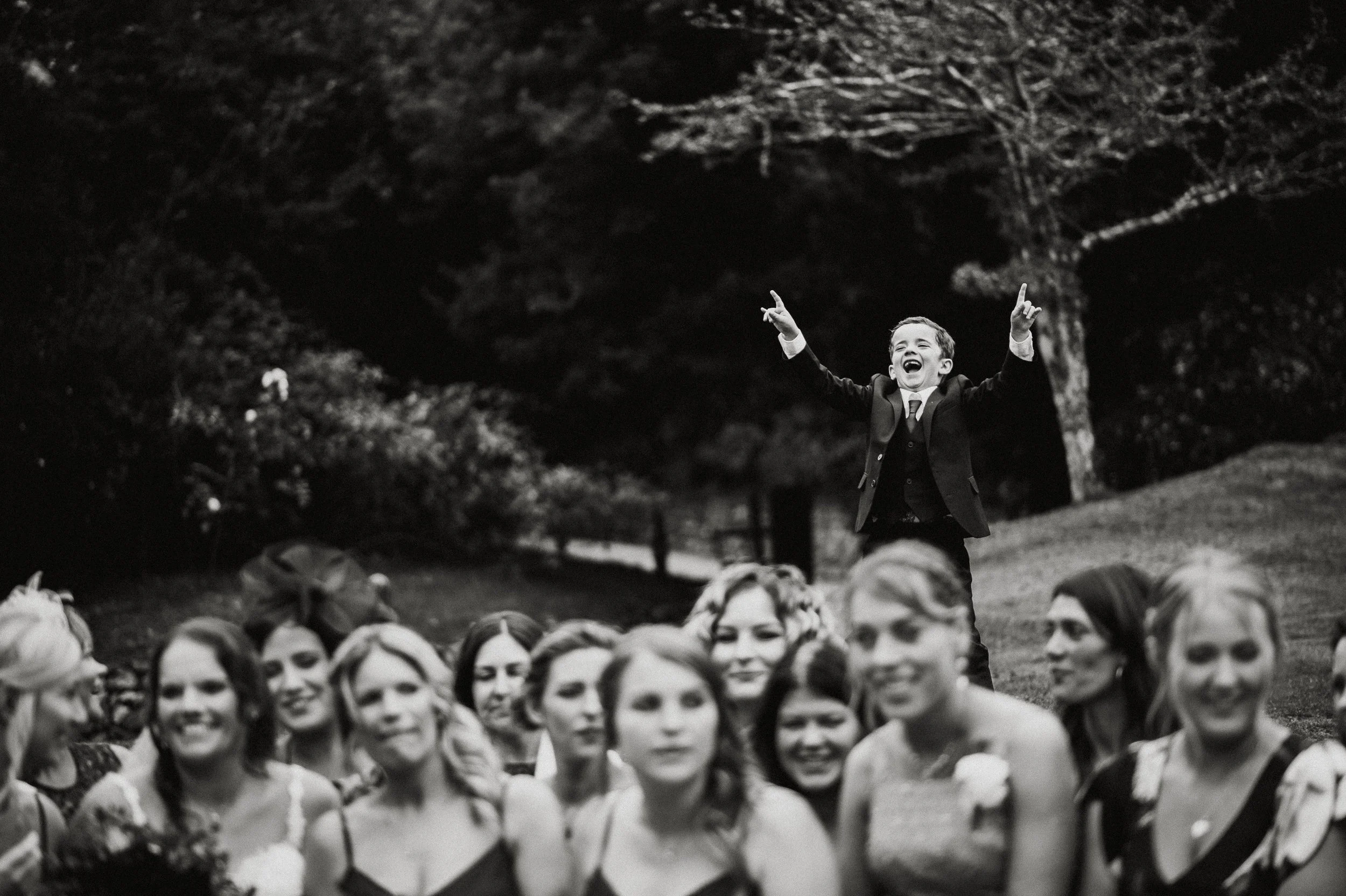 A boy in a suit, standing on a grassy hill, joyfully celebrating with arms raised at a wedding ceremony surrounded by women, with trees in the background.