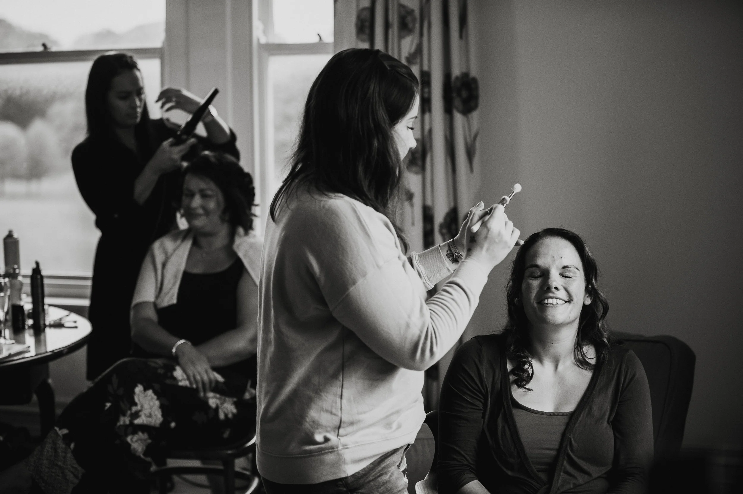 A woman receives makeup application from a makeup artist while another woman is seated in the background, smiling, in a well-lit room with windows and curtains.
